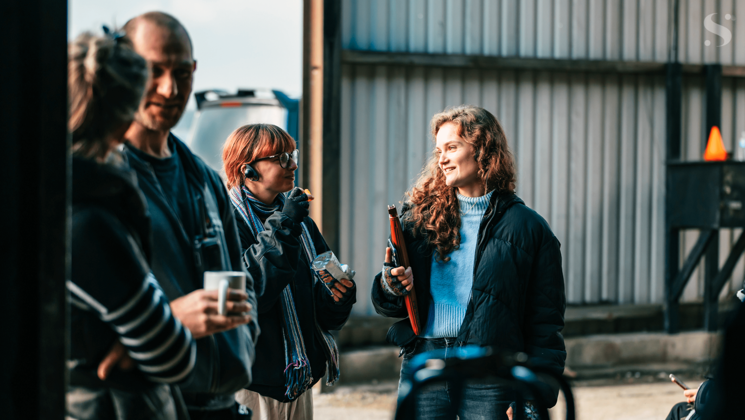 Hannah Alexander with crew on outdoor set near metal building for Singer Studios production, coffee cups in hand