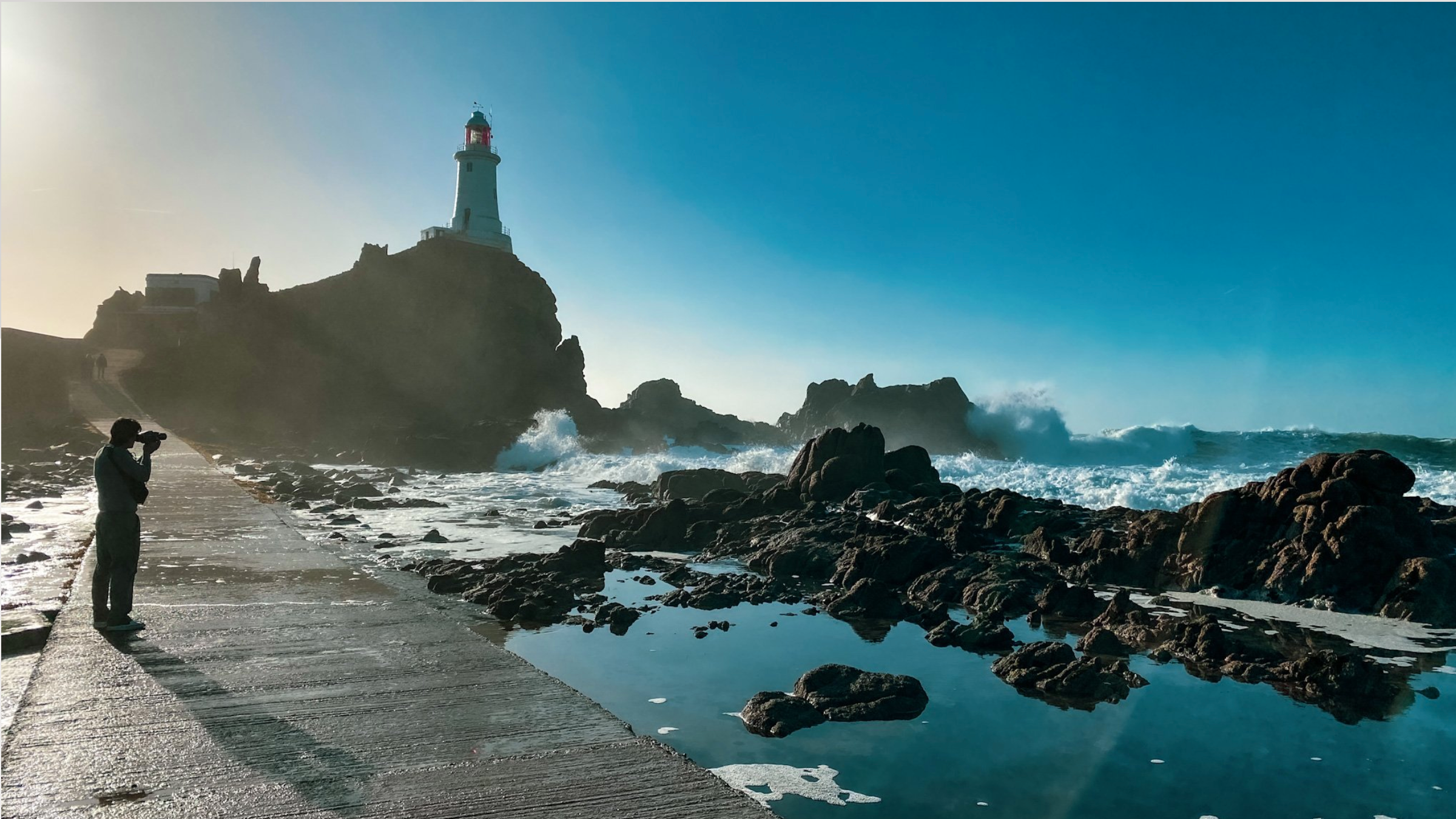 Cameraman Tim Cragg taking a photograph of a lighthouse on a rocky shoreline during sunset or sunrise, with waves crashing against the rocks for Singer Studios production, directed by Eloise Singer.