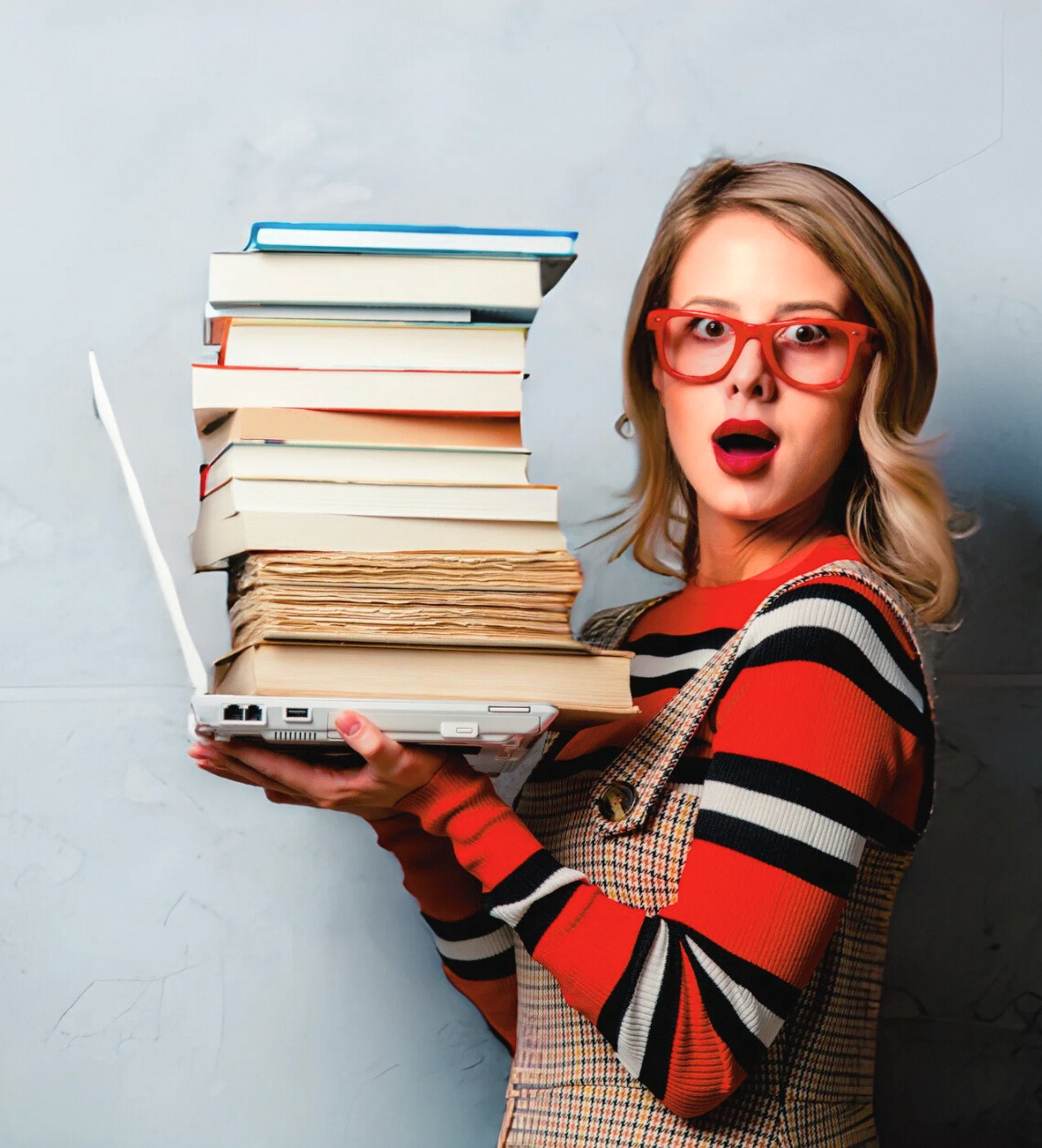 A woman with red glasses and red lipstick holding a laptop with a large stack of books and papers on top, surprised expression.