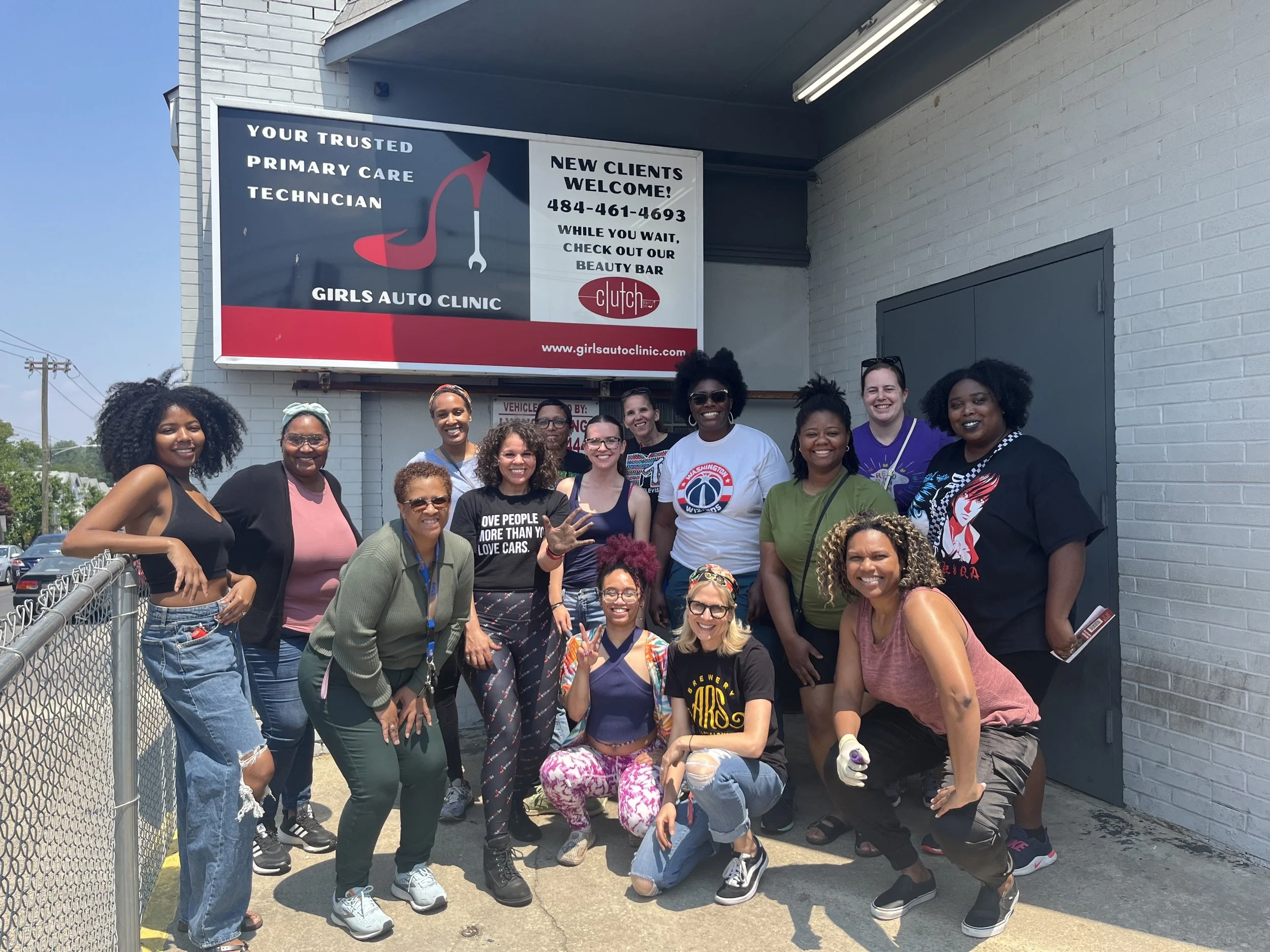 Group of women posing outside Girls Auto Clinic, smiling, some making peace signs, diverse in age and ethnicity, in front of a sign that reads 'Girls Auto Clinic' with a car logo.
