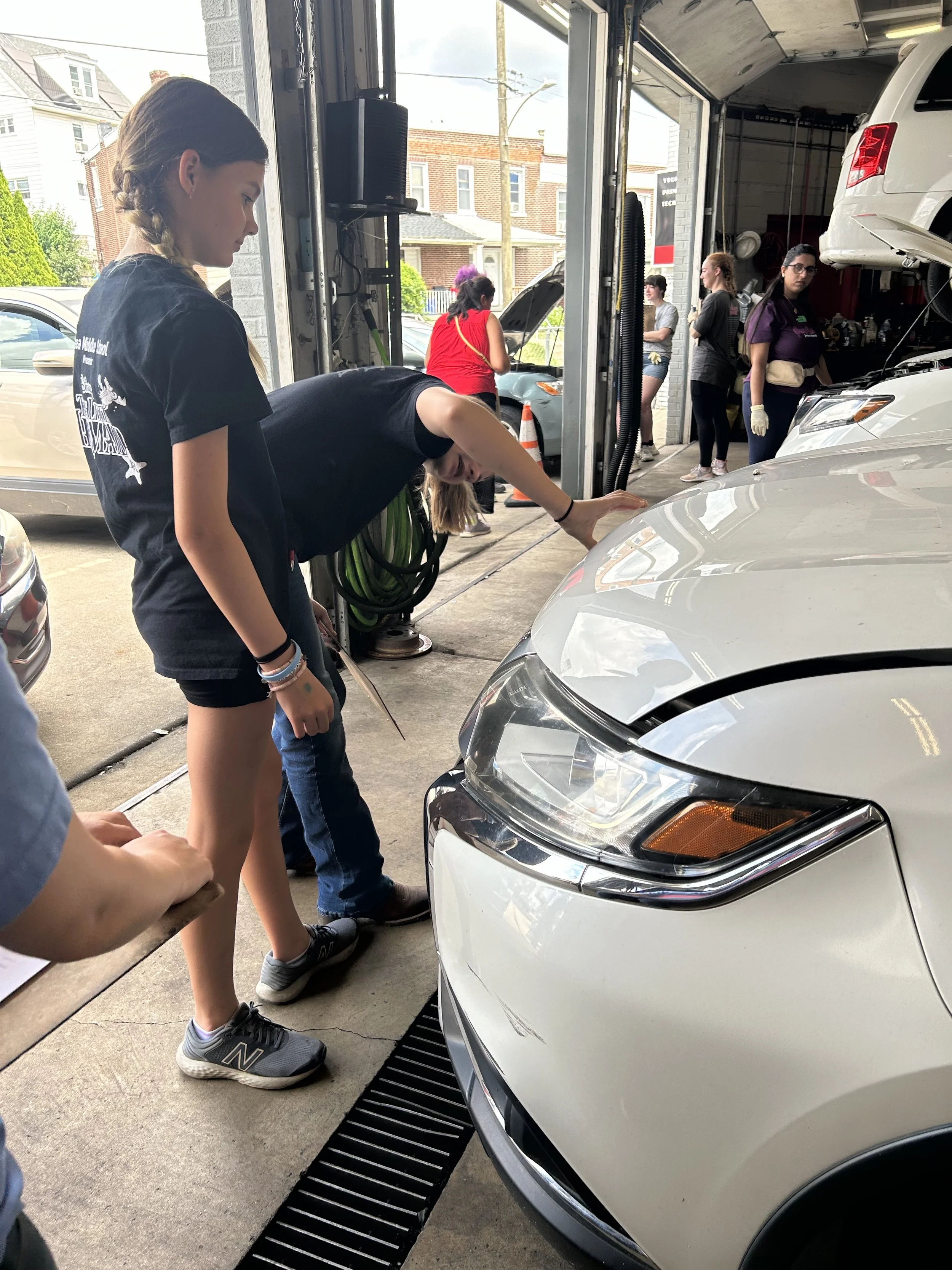 Multiple people working on cars inside an auto repair shop, with one person examining the front of a white car.