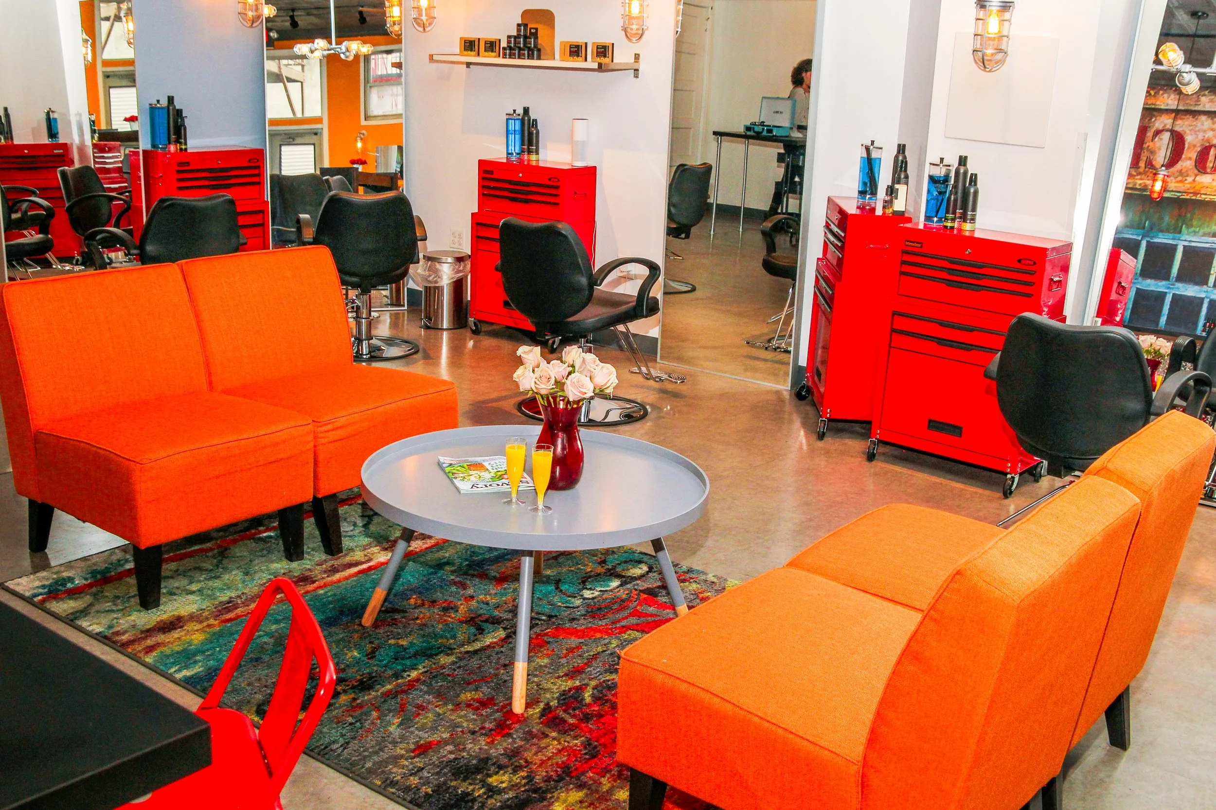 Stylish hair salon with black salon chairs, orange couches, red tool cabinets, and a gray coffee table with a red vase of pink roses and two yellow drinks. Modern lighting and large mirror reflect workspace.