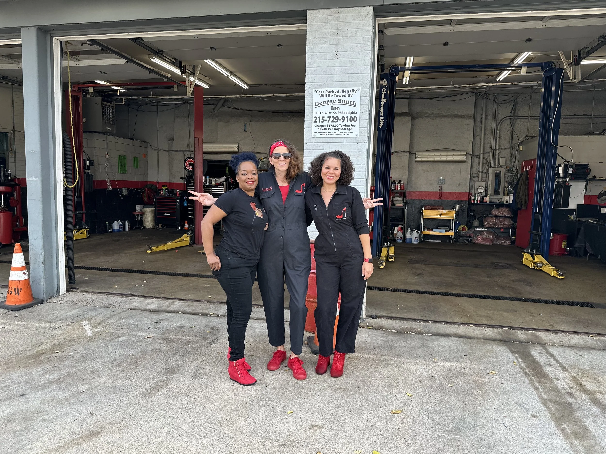 Three women standing in front of a car repair shop garage, smiling at the camera. They are wearing black uniforms with red shoes, and two of them have their arms around each other. The garage is open, showing car lifts, tools, and equipment inside.