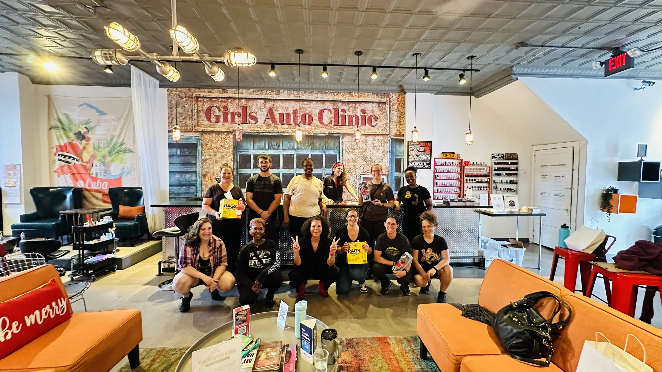 Group of people standing and sitting inside a colorful cafe with a sign that reads 'Girls Auto Clinic' in the background.