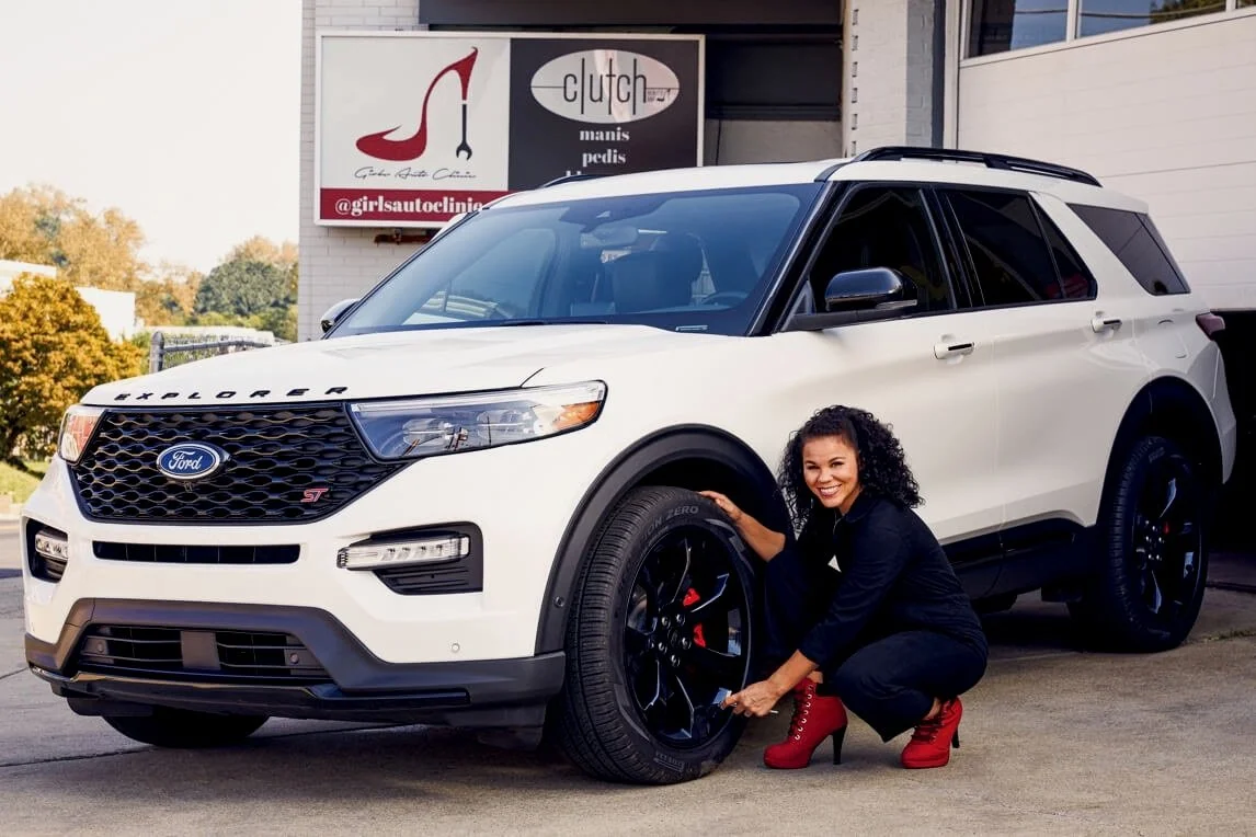 A woman with curly hair in a black jumpsuit and red high-heeled boots crouches next to a white Ford Explorer ST SUV, pointing at the tire on the driver's side, outside a building with a sign for a shoe and pedicure business.