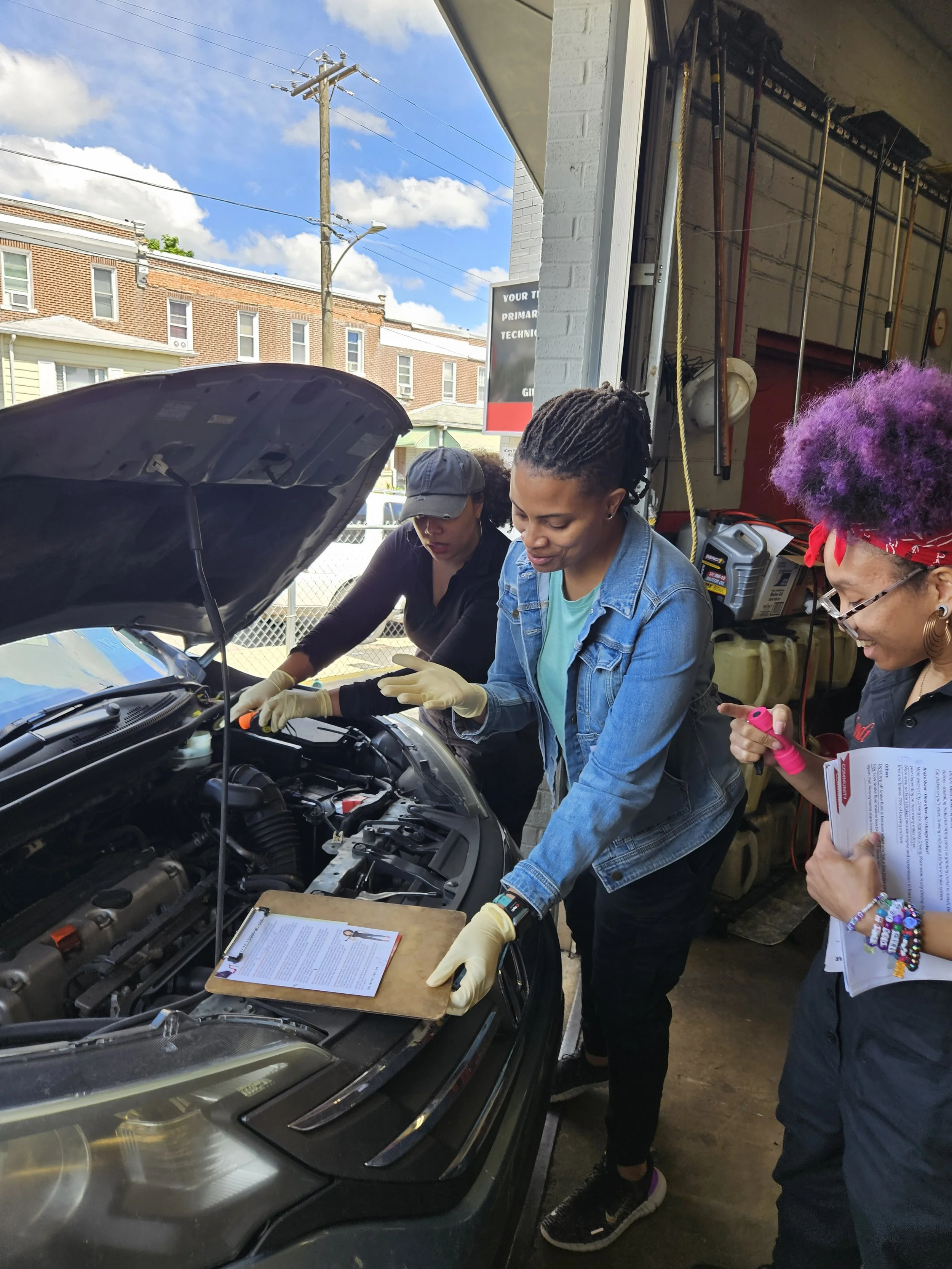 Three women in a garage working on a car's engine, with one woman holding papers and a highlighter, and another woman writing on a clipboard, under a sunny sky.