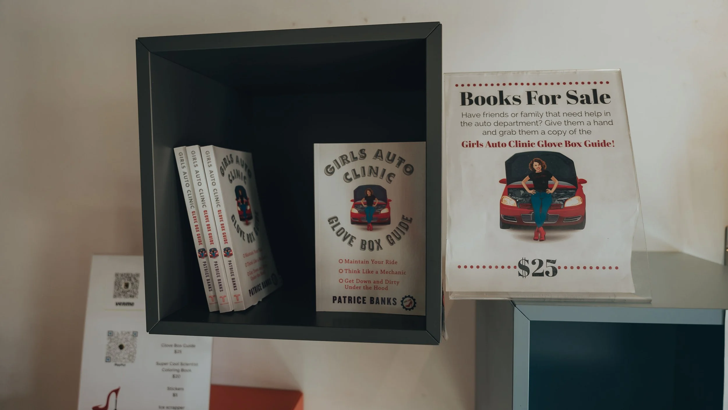 A shelf displays copies of a book titled 'Girls Auto Clinic Glove Box Guide' and a promotional sign advertising the book for $25. The sign features an image of a woman leaning against a red car with an open hood.