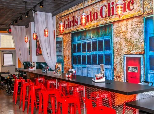 Interior of a restaurant with a long black bar counter and high red chairs. Behind the bar is a colorful mural of a rustic building with a sign that reads 'Girls Auto Clinic.' There are hanging lantern-style lights and white curtains dividing the spa