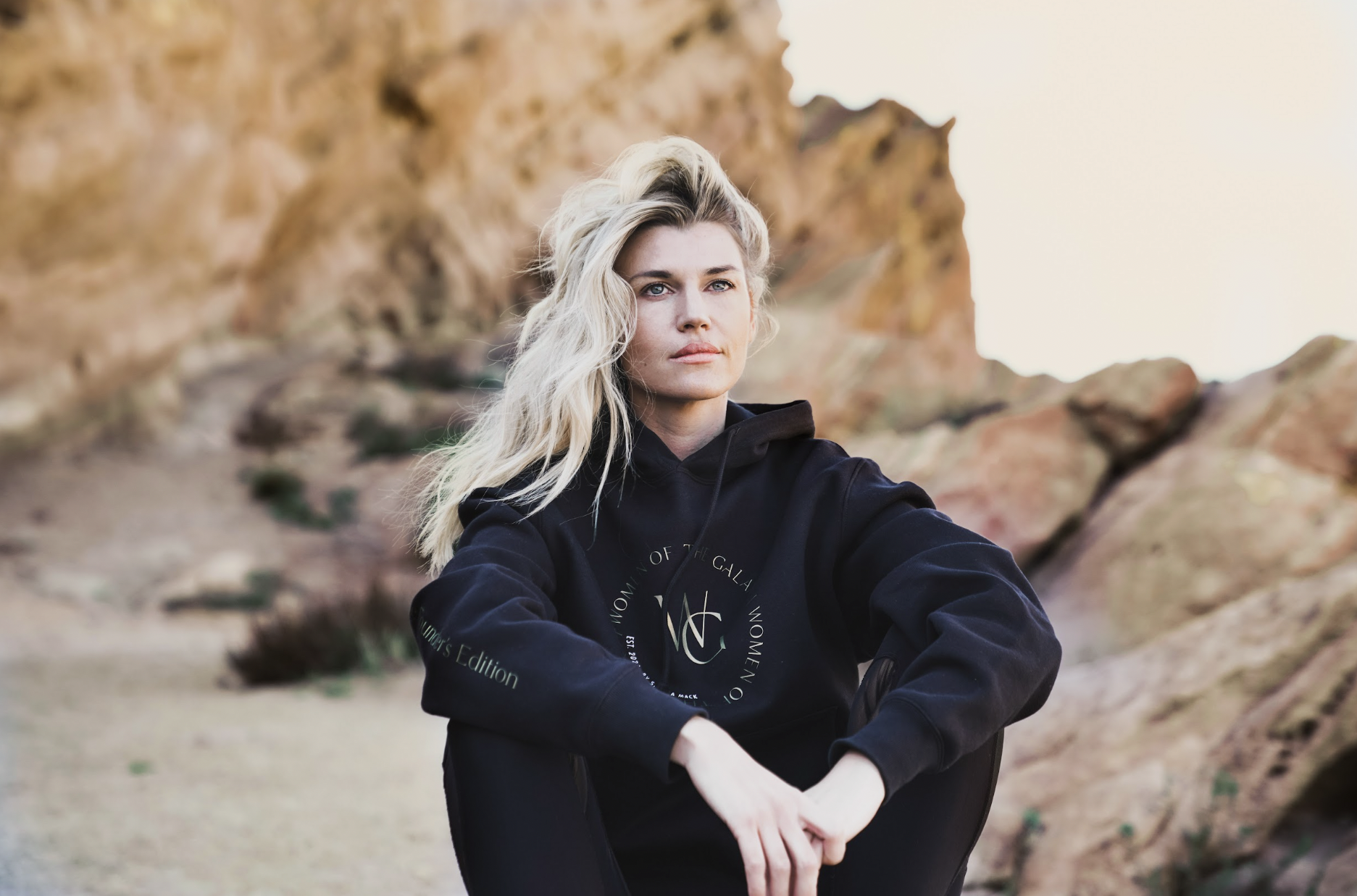 Woman in black hoodie seated on coastal rocks — apparel product photographer San Diego