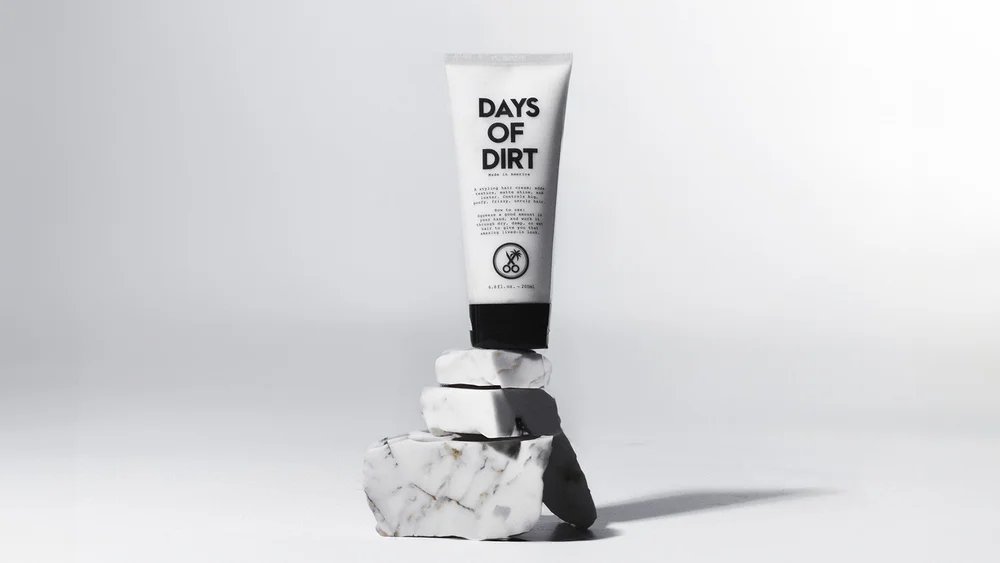 Minimalist commercial skincare photograph of a Days of Dirt tube balanced on textured stone blocks, sculpted by hard directional side light that creates a long graphic shadow while preserving clean packaging edges and readable typography