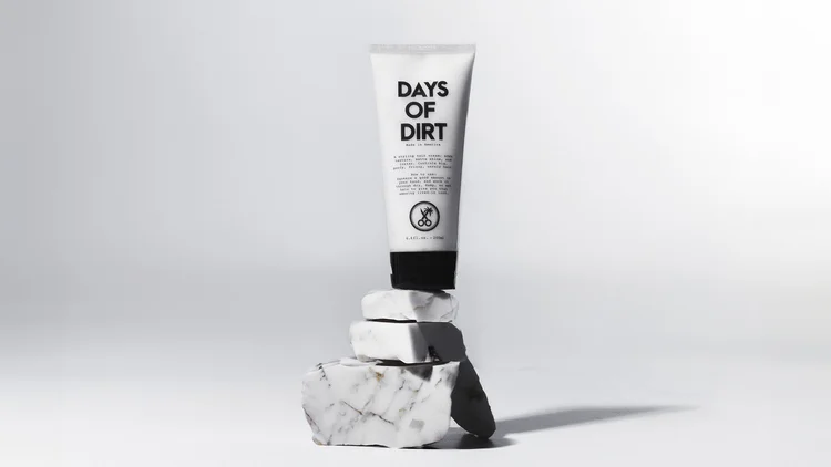Minimalist commercial skincare photograph of a Days of Dirt tube balanced on textured stone blocks, sculpted by hard directional side light that creates a long graphic shadow while preserving clean packaging edges and readable typography