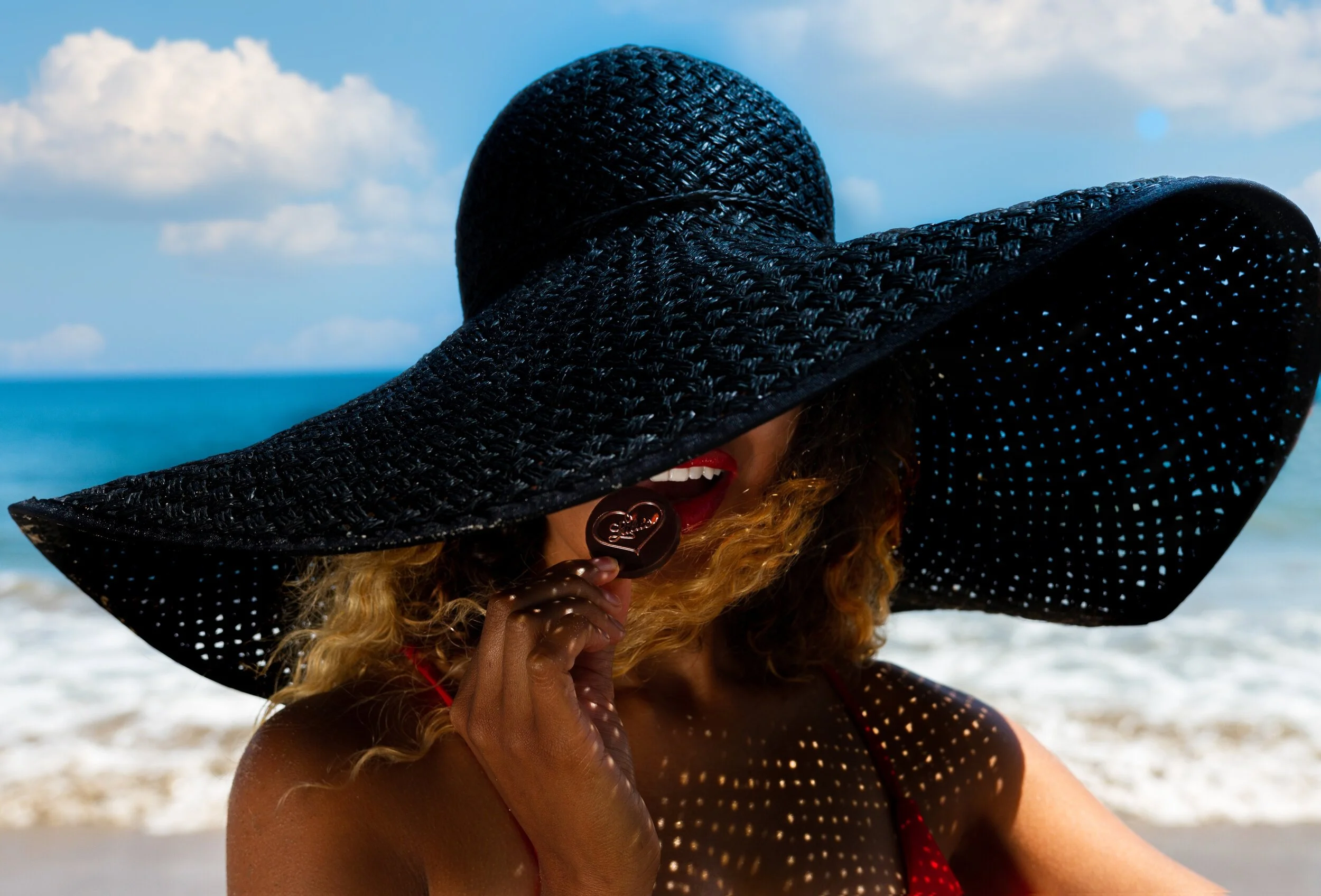 Accessories product photograph of an oversized black woven sun hat worn by a model at the beach, photographed in bright coastal sunlight with the ocean in the background