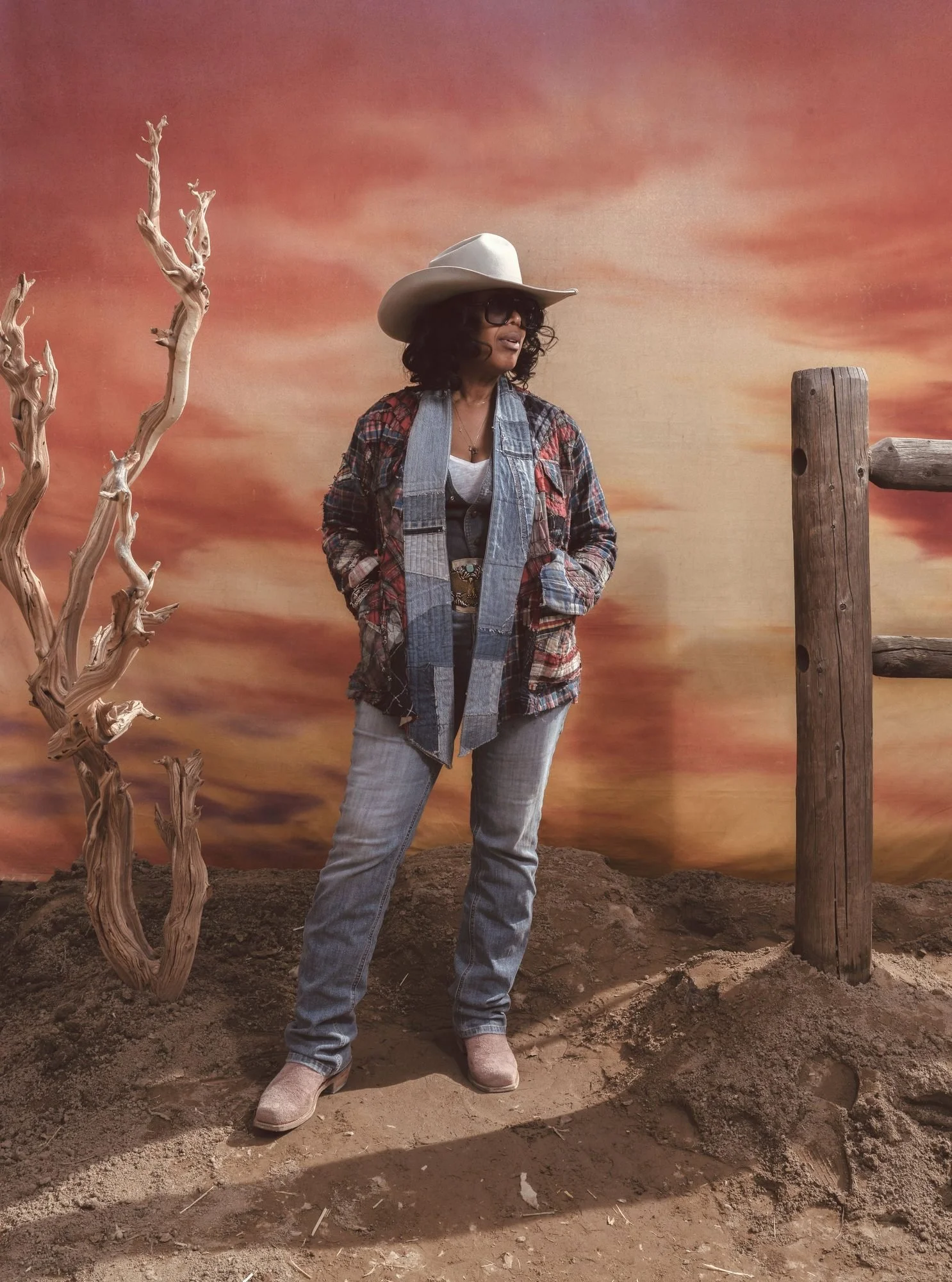 Guest in a white cowboy hat and patchwork denim jacket standing by a fence on the sunset set, Iron Rodeo portrait studio