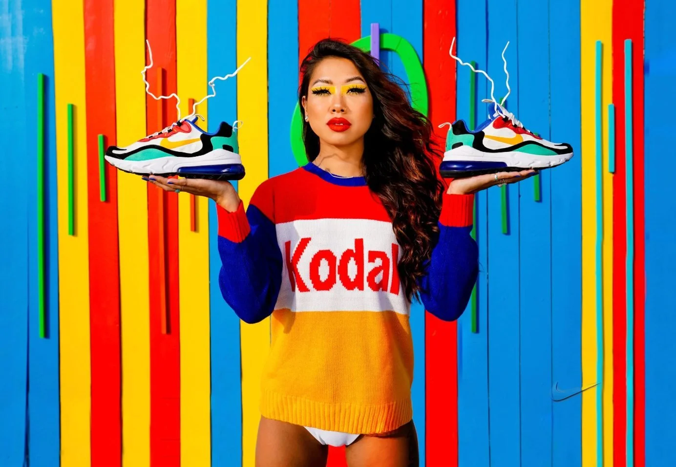 Woman holding Nike sneakers against a saturated red, yellow and blue mural, using symmetrical composition and bold graphic color blocking.