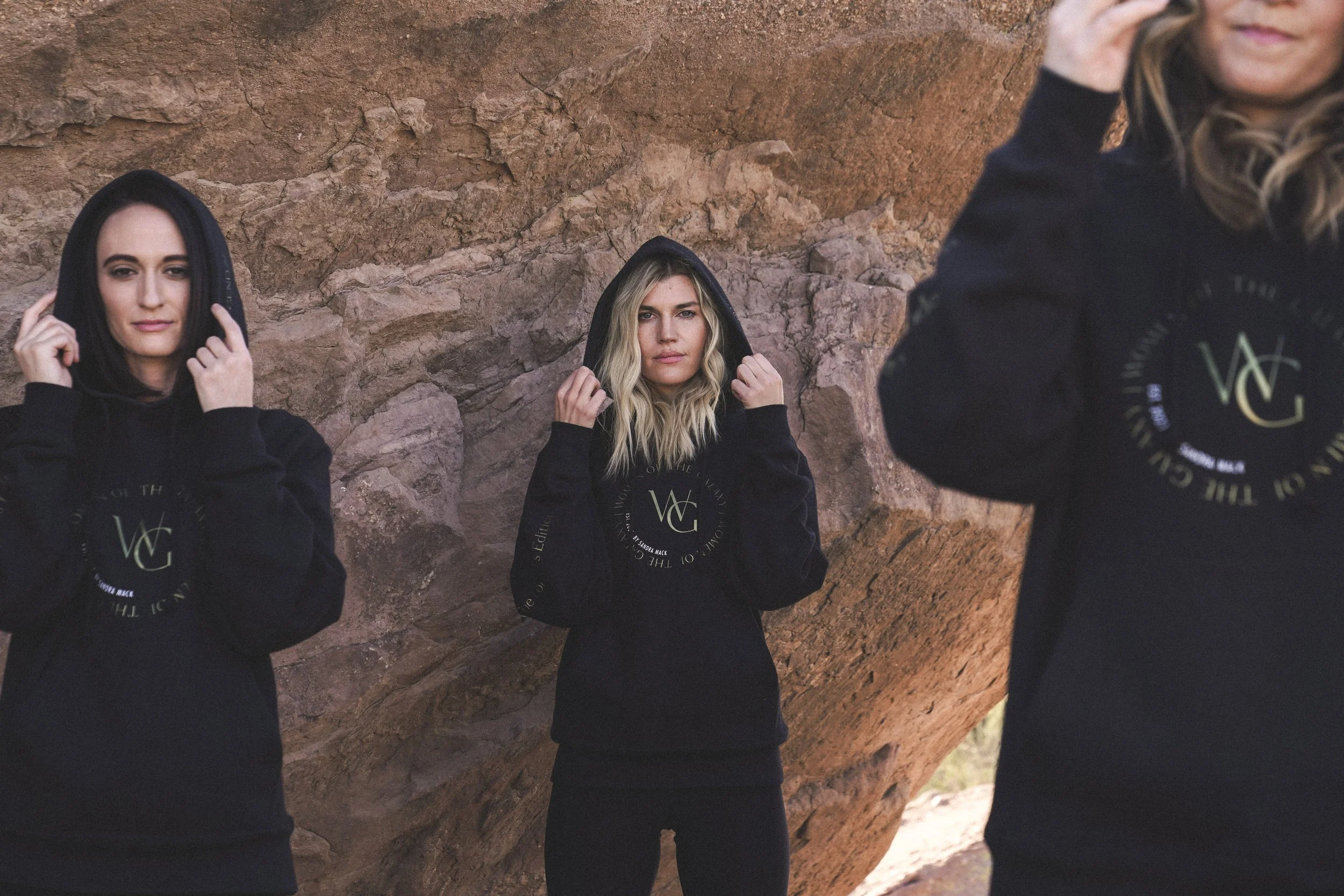 On-model black hoodie product photography using soft directional light and a textured stone setting to retain shadow detail in the dark fabric, define hood volume and silhouette, and keep subtle branding visible without flattening the garment