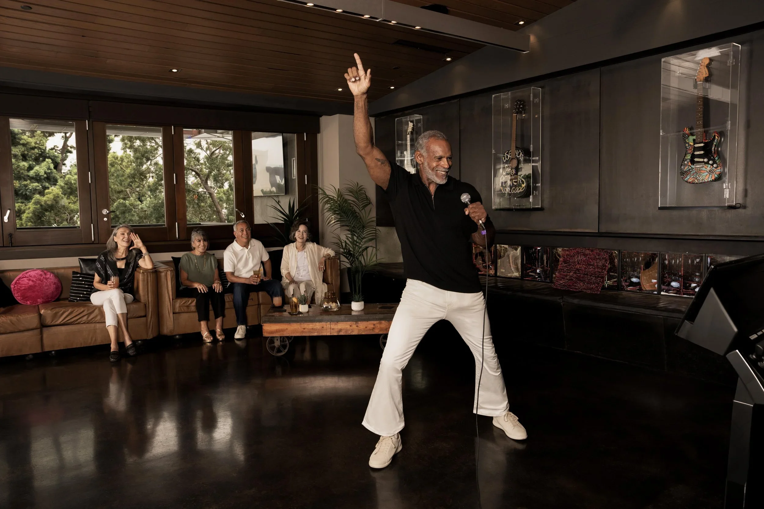 Cinematic commercial campaign recreating an iconic movie-inspired karaoke party scene, with a performer entertaining seated guests in a modern interior, photographed with controlled directional light, layered composition and carefully balanced shadow