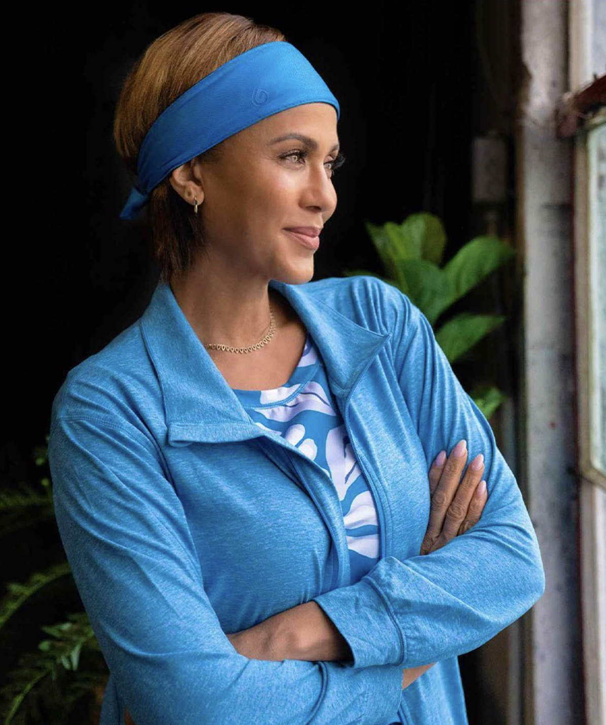 A woman with short brown hair, wearing a blue sports headband and matching blue athletic jacket, stands with arms crossed indoors near a window, looking to her right with a contemplative expression.