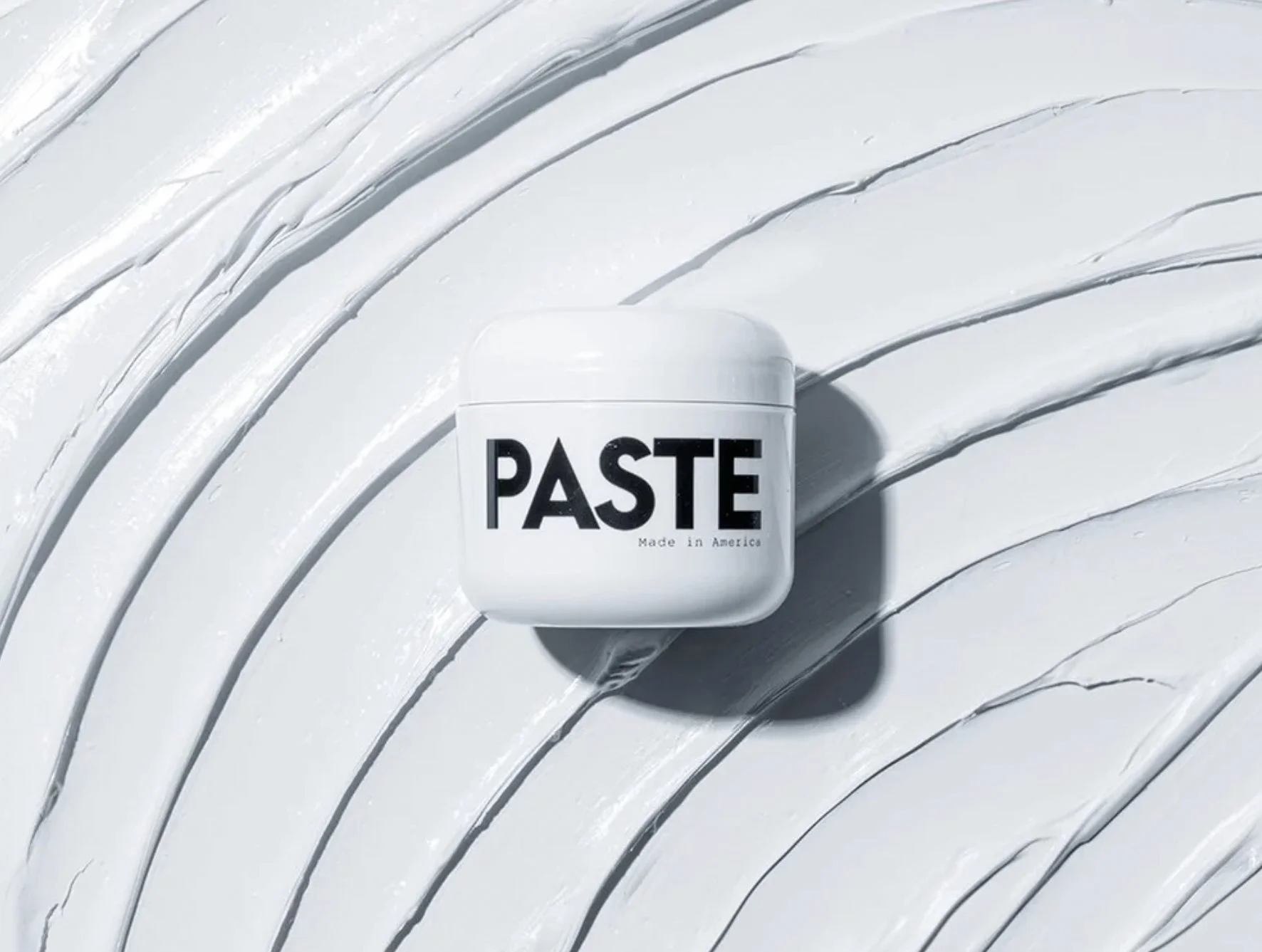 Days of Dirt Paste jar on a sculpted white surface with hard light and graphic shadows, men's grooming product photography