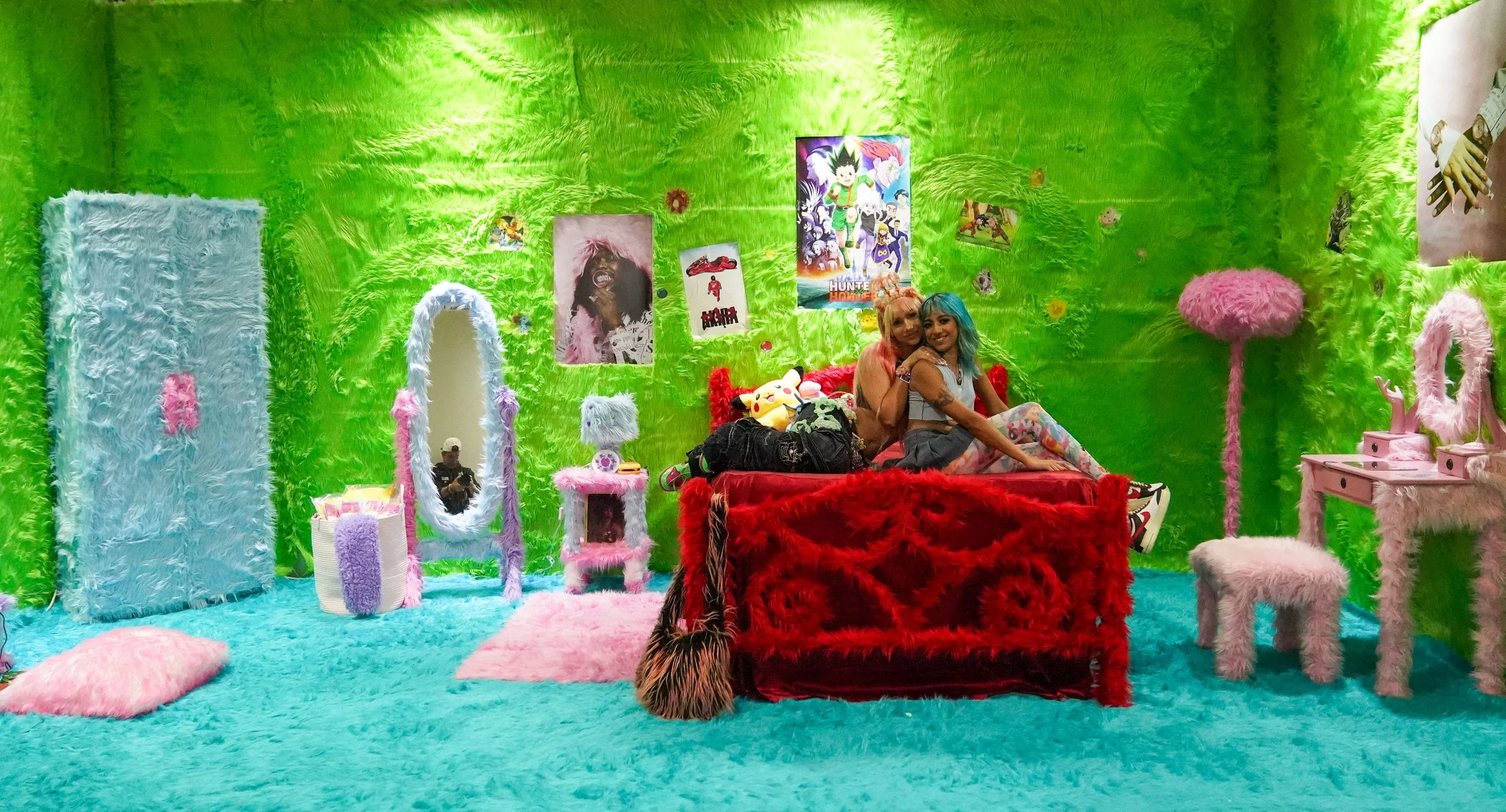 Two guests posing on a red fur-covered bed inside an immersive neon-green installation, surrounded by pastel furniture, anime artwork and playful pop-culture details