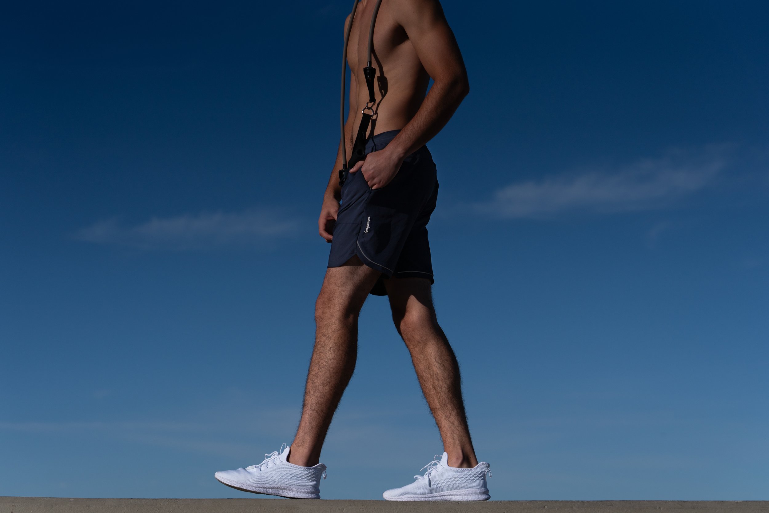 A shirtless man wearing dark shorts and white sneakers walking outdoors against a clear blue sky.