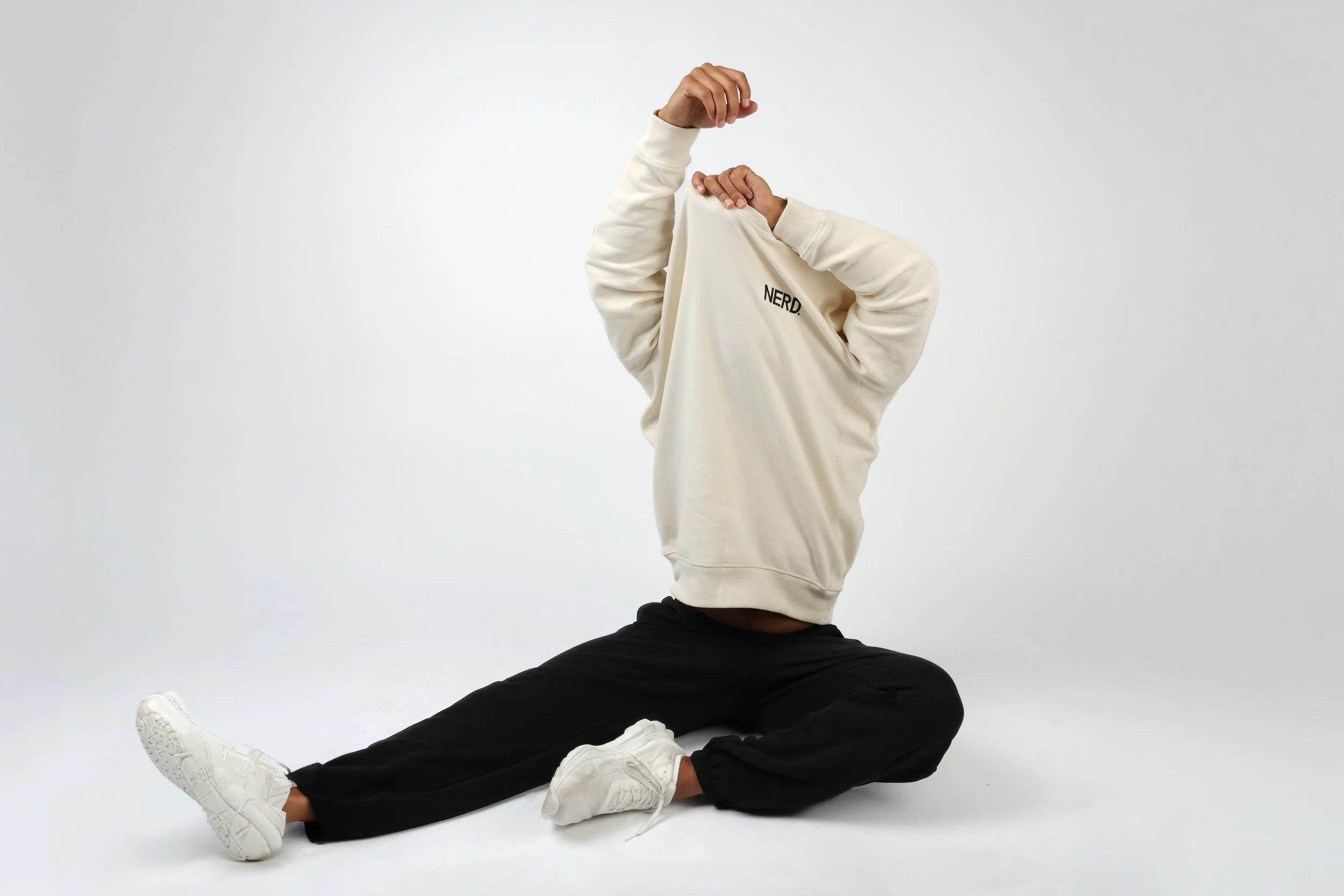 Seated model wearing a cream NERD sweatshirt in a clean white studio product photograph