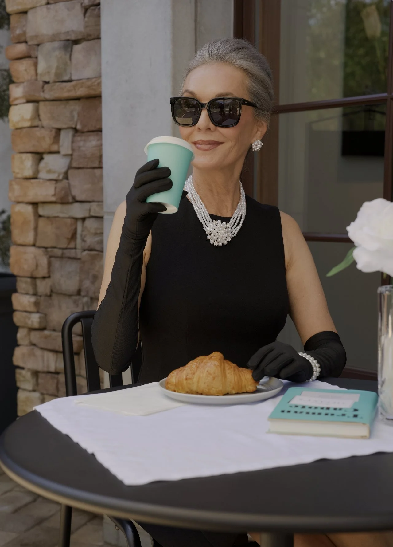 Silver-haired model in a black dress, long gloves and pearls sipping coffee at a cafe table, a Breakfast at Tiffany's scene shot for TRAFFIK and Alignment Health