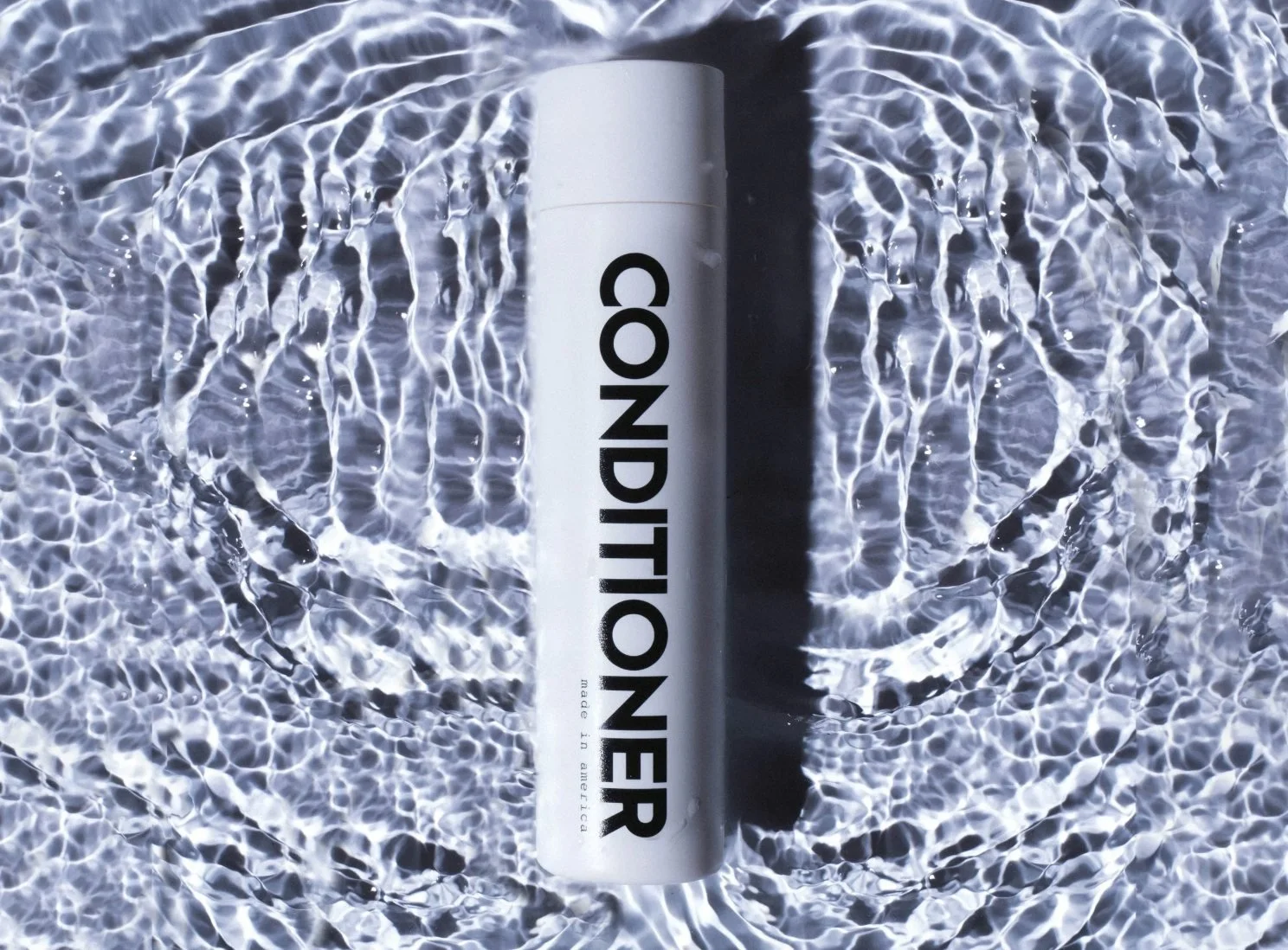 White Days of Dirt conditioner bottle in rippling water with high-contrast studio light, hair care product photography