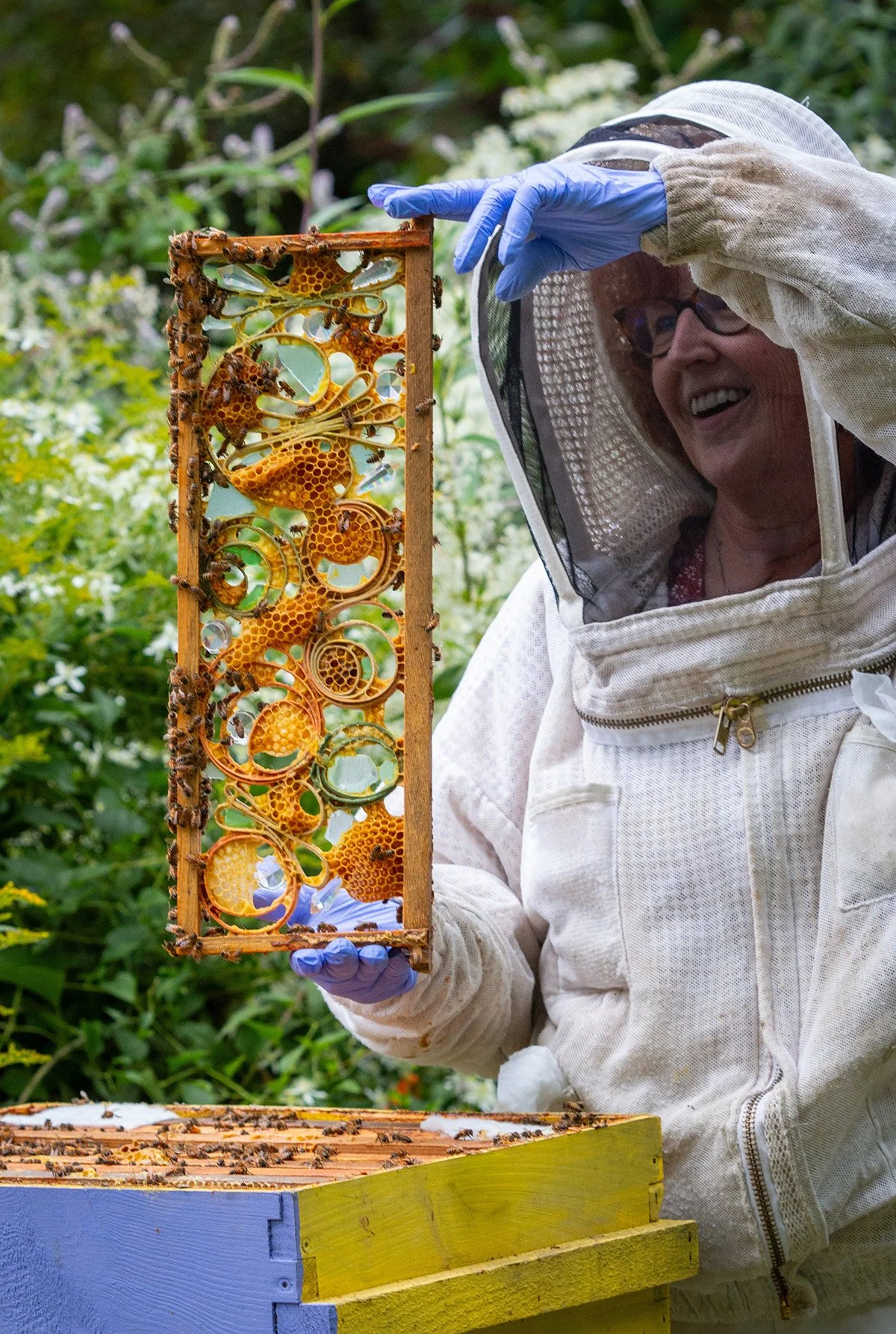 Susan Medyn wearing a beekeeper's suit, holding up a piece of her honeycomb artwork over the beehive