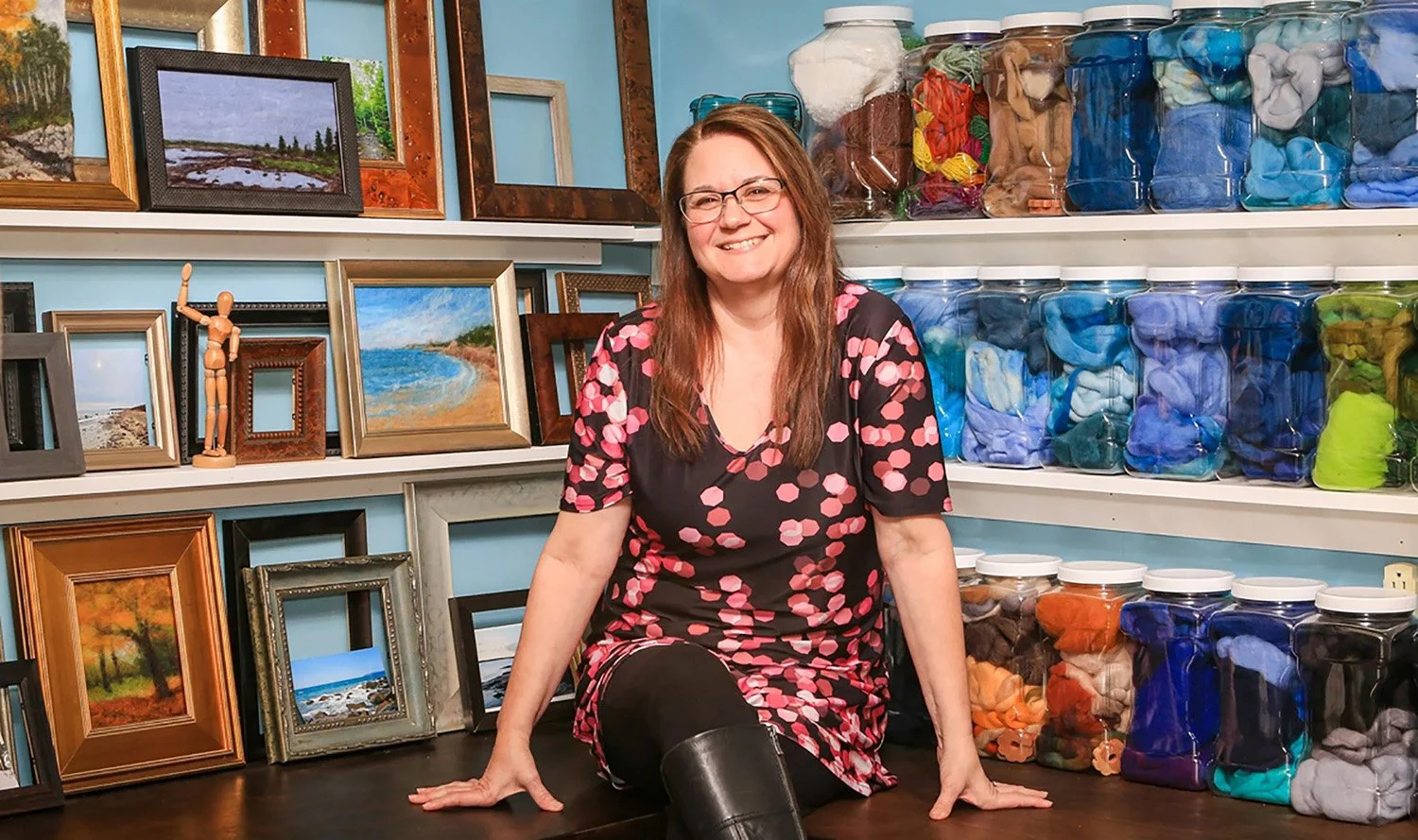 Stephanie Stroud seated with finished artwork, frames, and canisters of yarn behind her