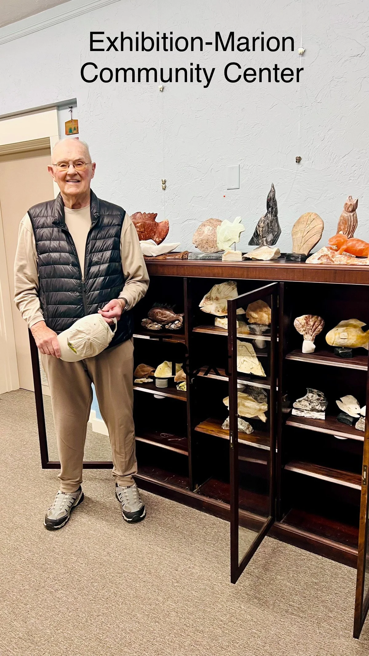 Hugh Kelly standing next to a case of sculpture. A caption atop the photo reads "Exhibition: Marion Community Center"