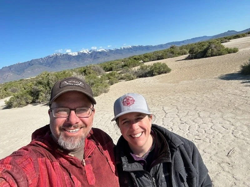 Two smiling people taking a selfie in the Alvord desert with mountains and snow-capped peaks in the background.