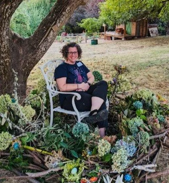 A woman sitting on a white garden bench under a tree surrounded by colorful flowers and plants in a garden.