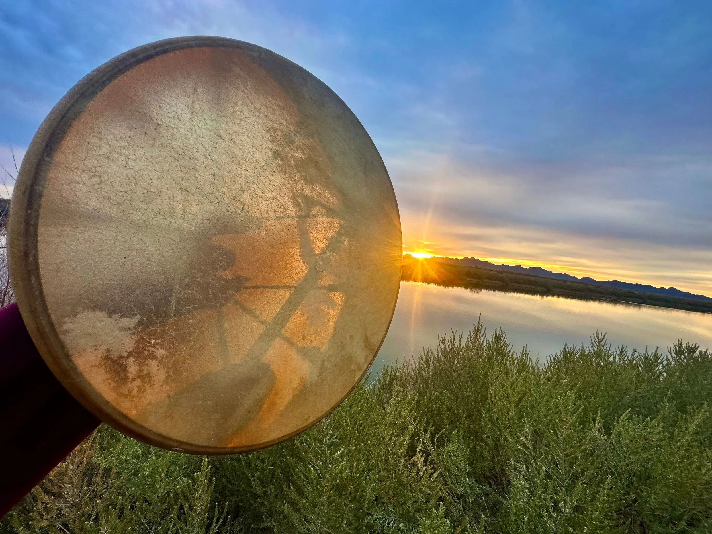 A hand holding a round hand drum, in front of a sunset over a body of water with mountains in the distance and bushes in the foreground.