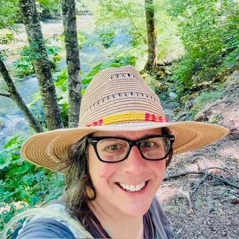 Smiling woman in a straw hat with black glasses in a forest near a river.