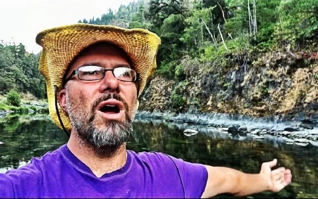 Man wearing a straw hat and glasses taking a selfie outdoors by a river, with forested hills in the background.