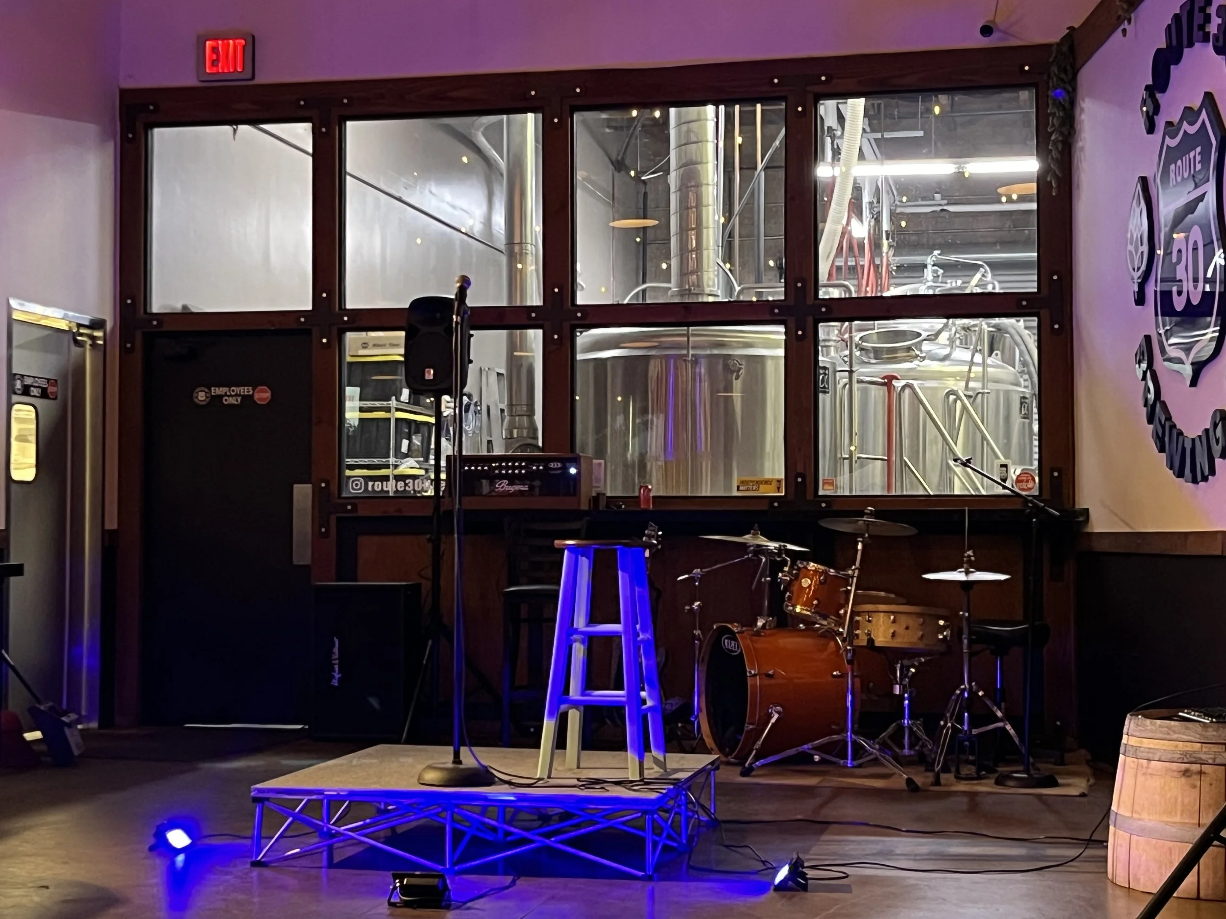 Empty music stage with a drum set, microphone, and stool, in front of a large window showing brewing tanks in a brewery interior.