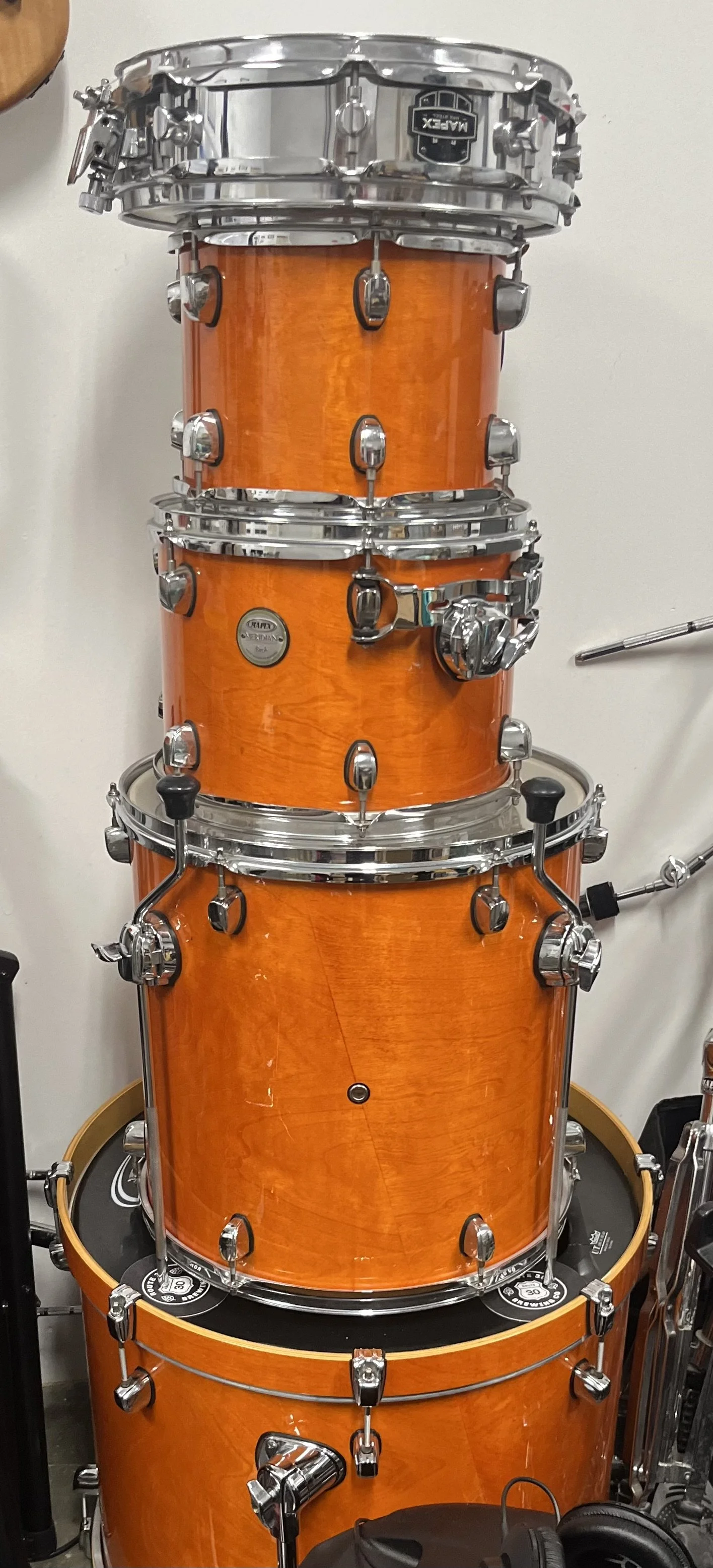 Stacked orange drum set drums with chrome hardware and bass drum on the bottom, against a white wall.