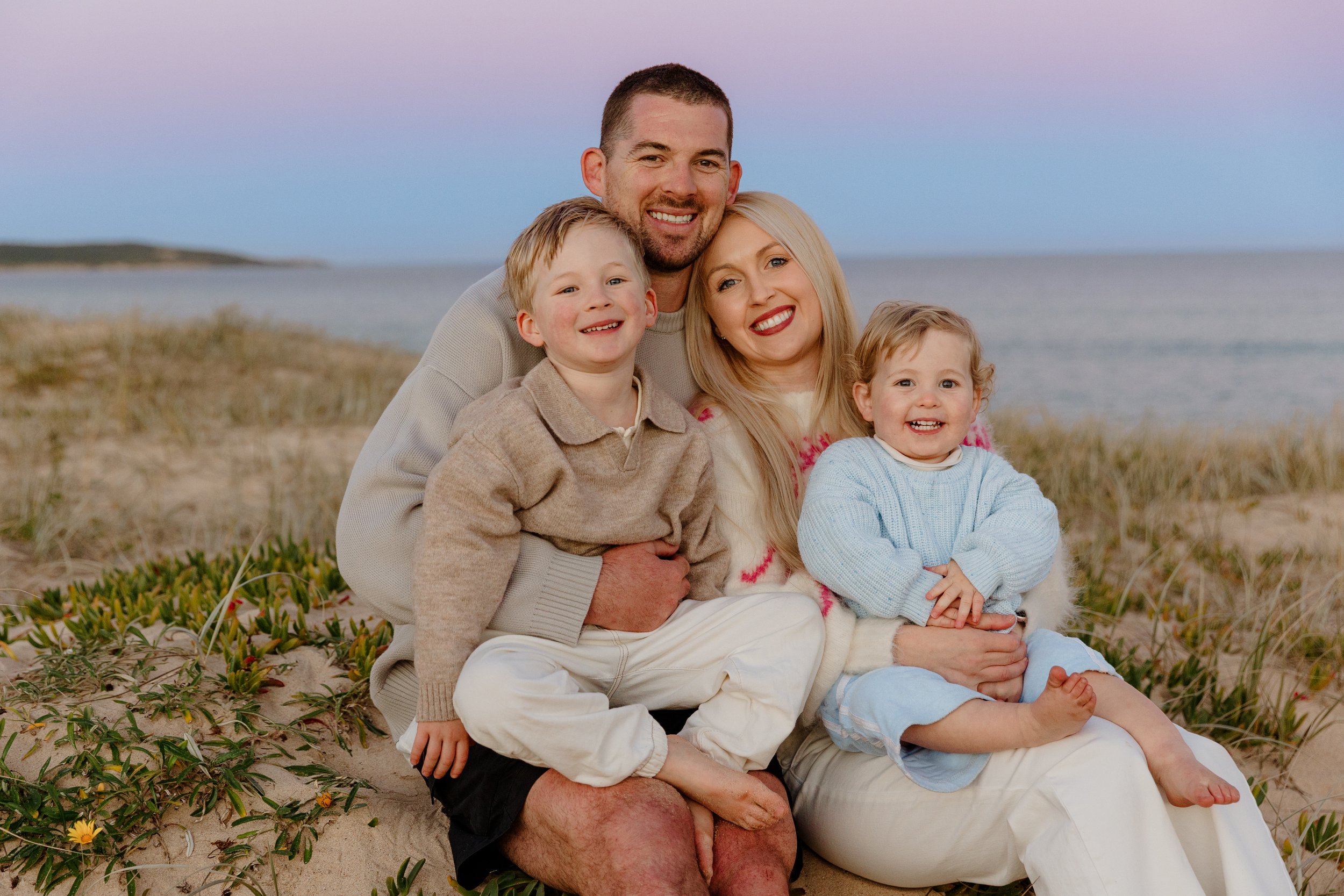 A family of four sitting on the sand at a beach during sunset, smiling and hugging each other.
