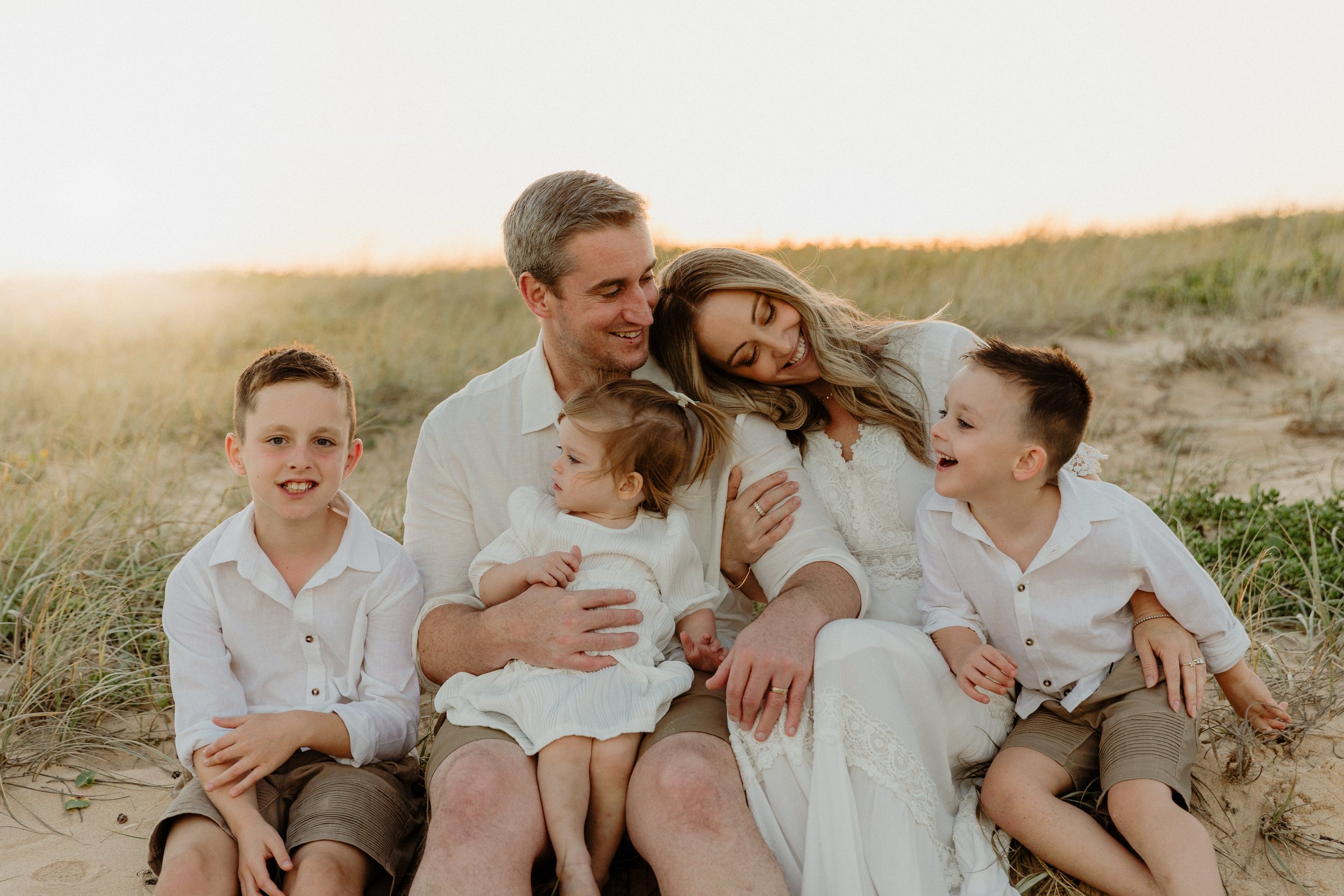A family of six, including two adults and four children, sitting on sand dunes with beach grass during sunset, smiling and enjoying together.