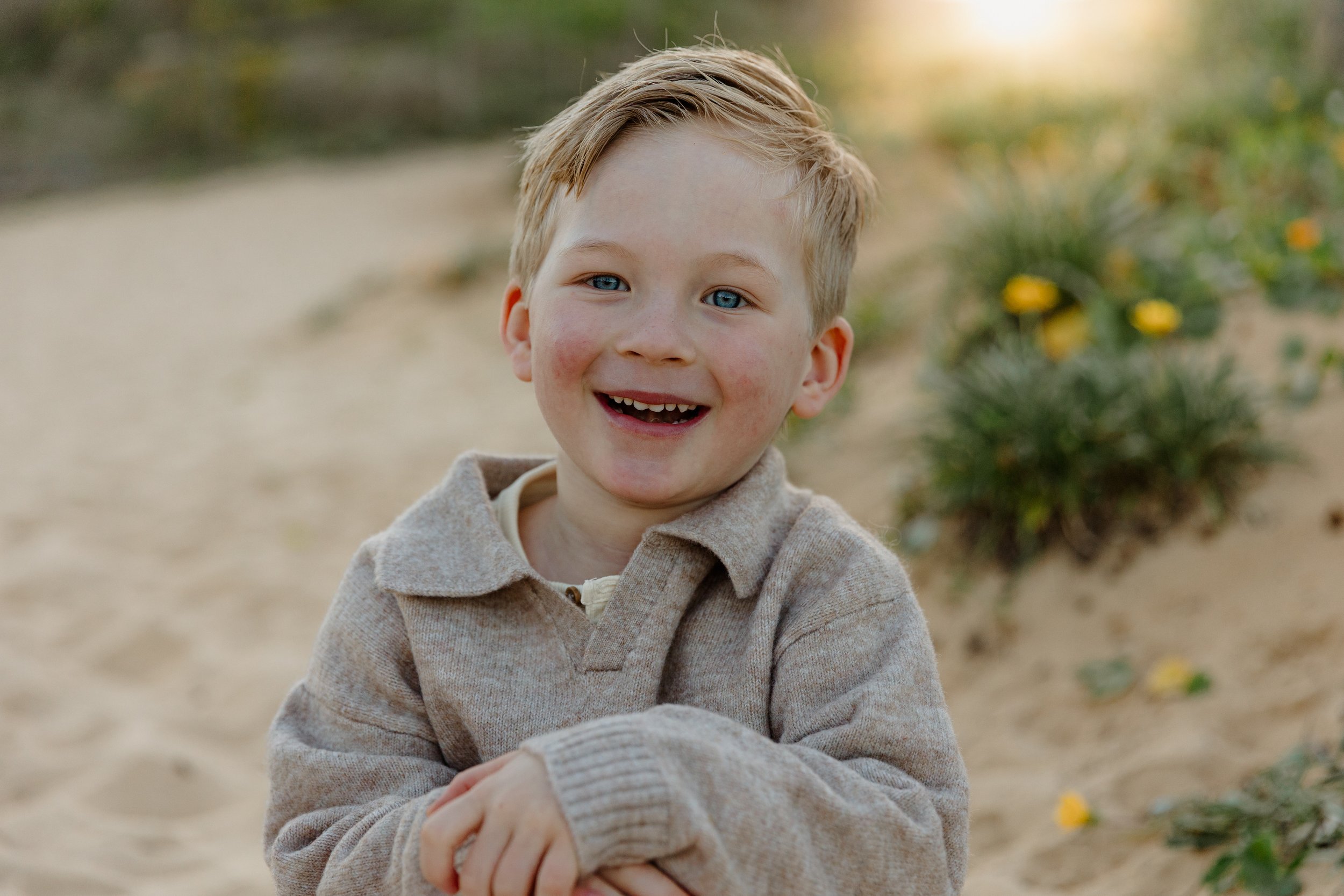A young boy with red hair and blue eyes smiling outdoors on a sandy path, wearing a beige sweater.