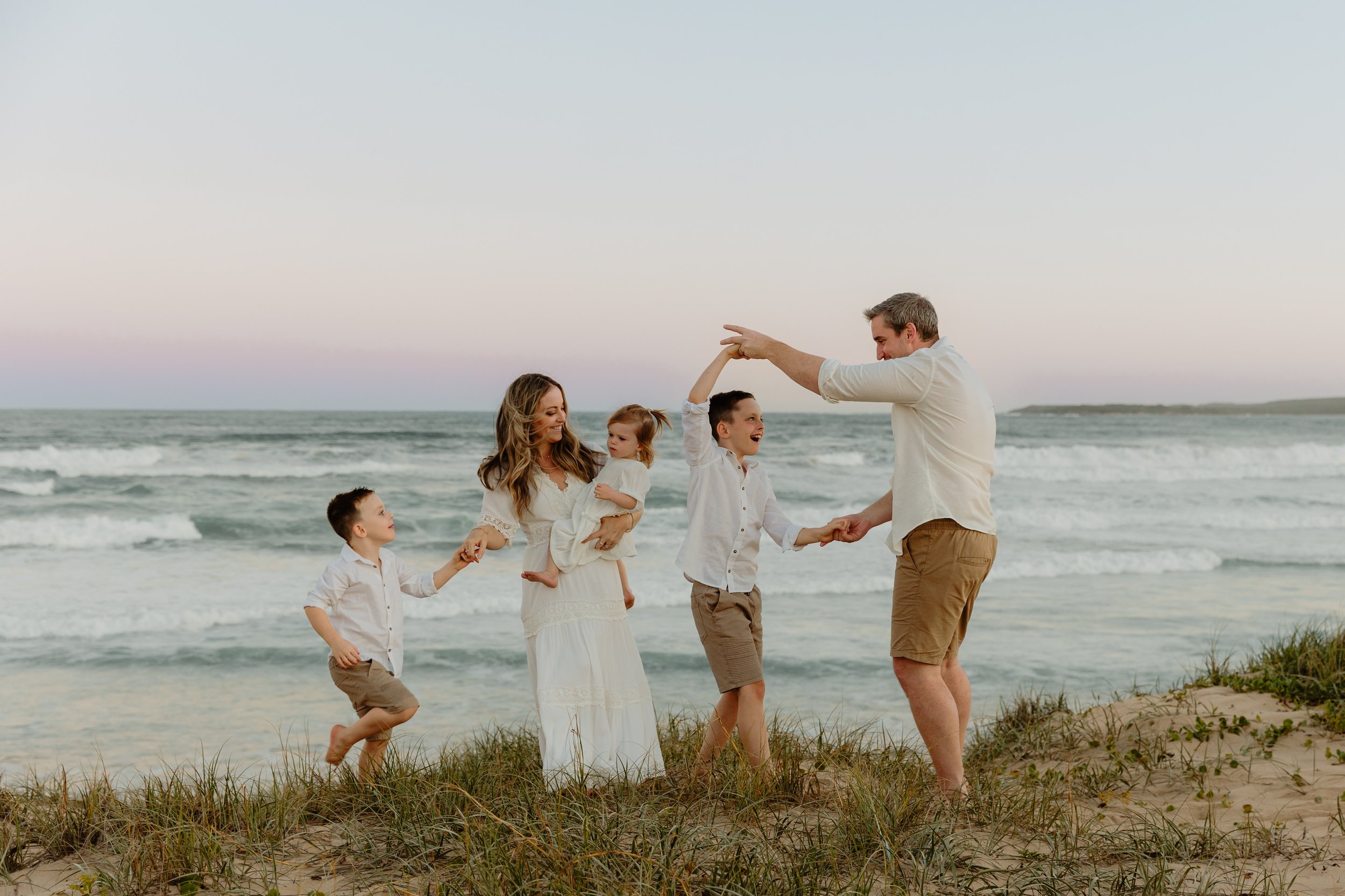 Family of five enjoying a playful moment on the beach, holding hands and dancing in the sand with ocean waves in the background.
