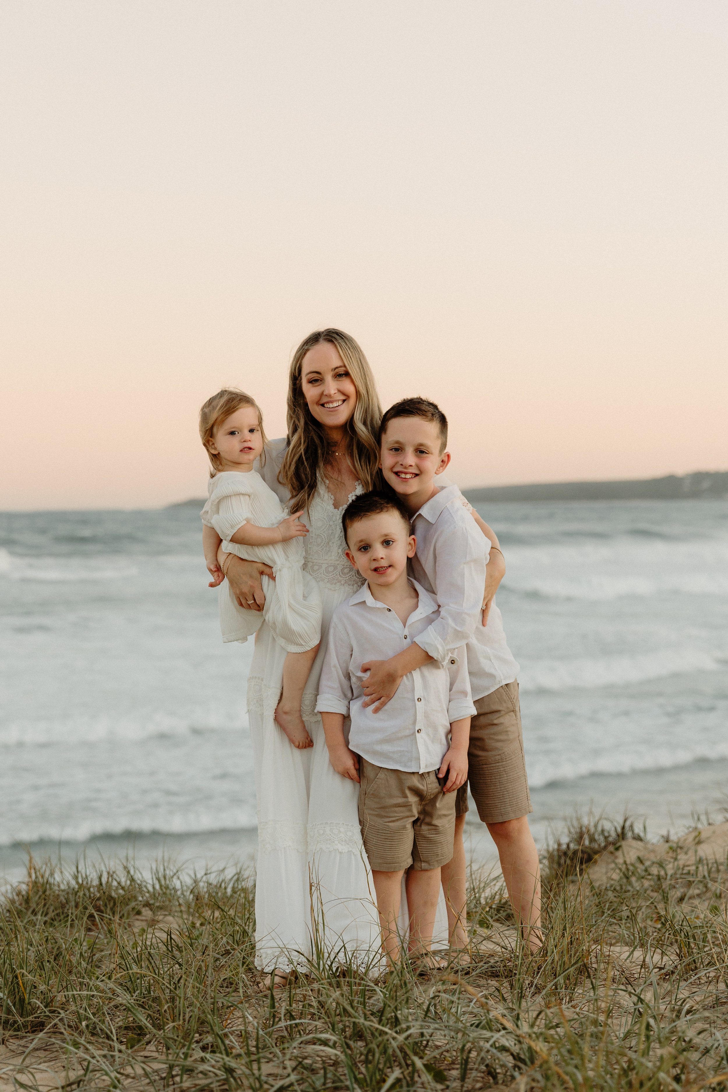 A woman and three children standing on the beach at sunset, smiling, with waves and sky in the background.