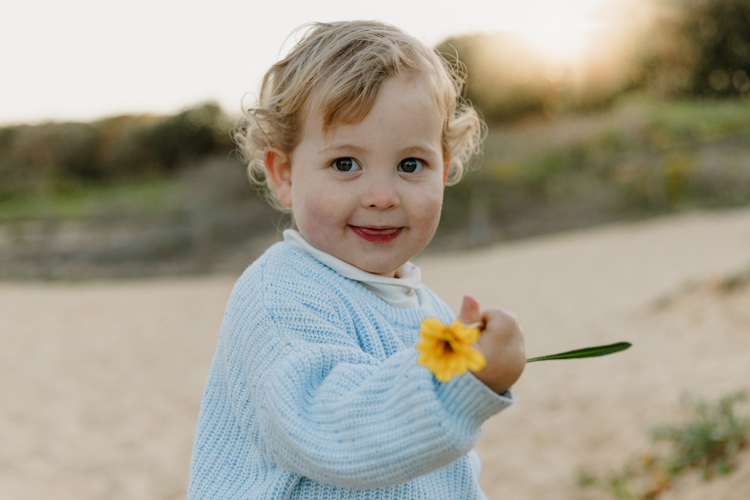 A young child with curly blonde hair and blue eyes smiling and holding a yellow flower in an outdoor setting during the daytime.