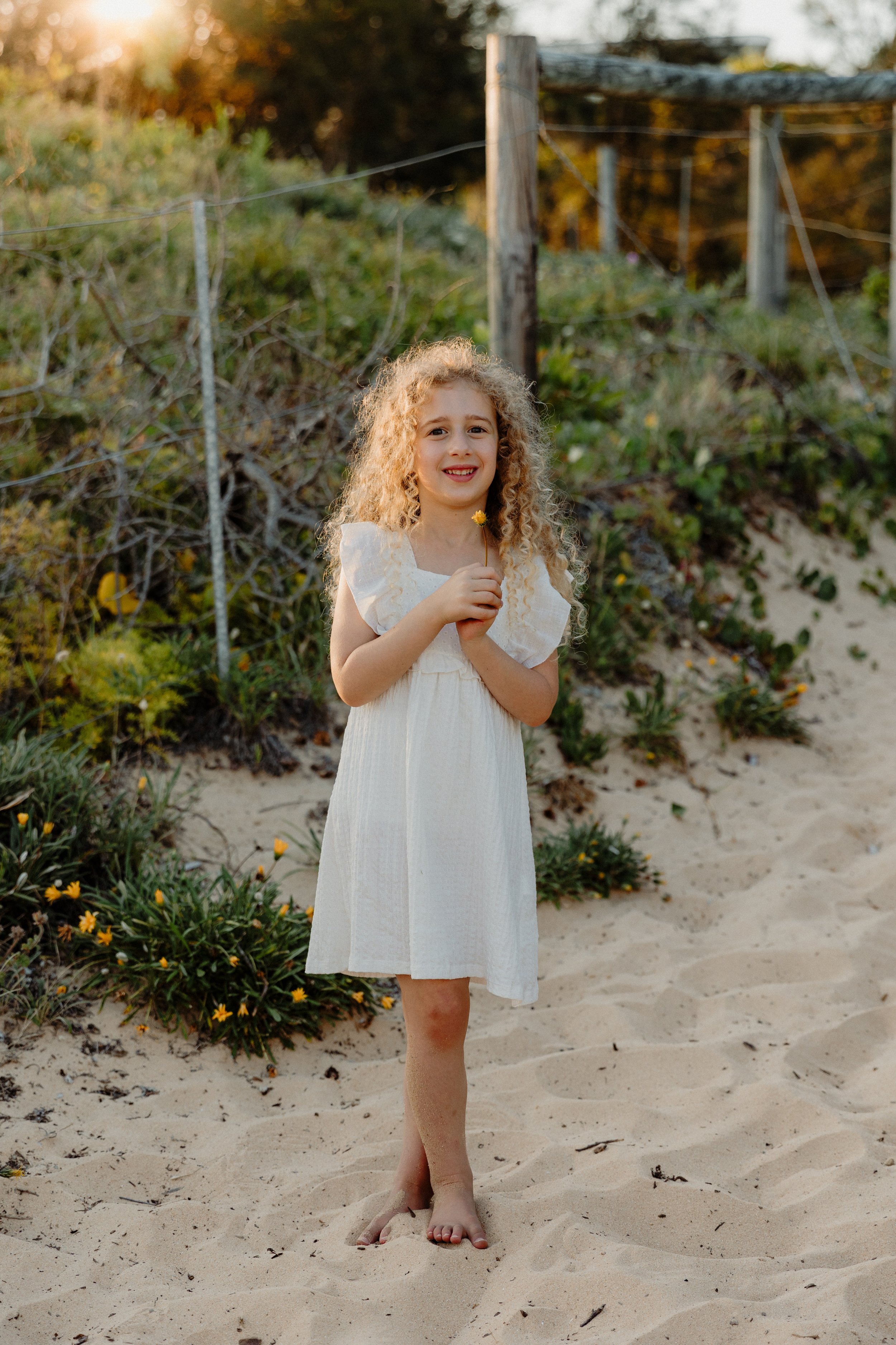 A young girl with curly blonde hair in a white dress standing barefoot on sandy ground, holding a small yellow flower, at a beach or coastal area during sunset.