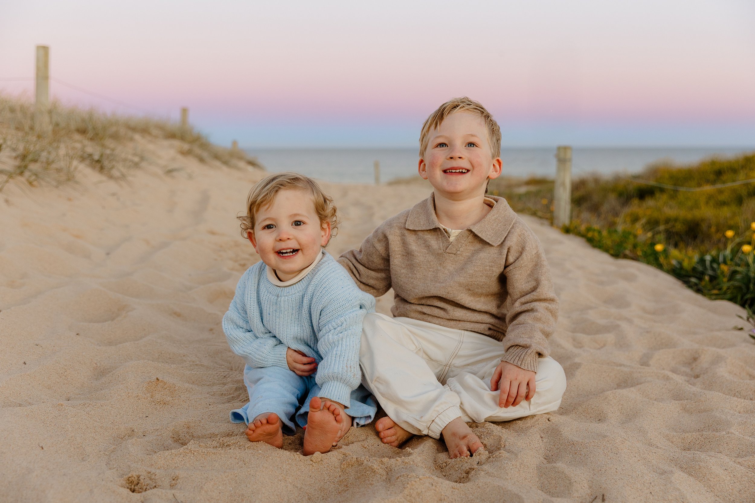 Two young children, a boy and a girl, sitting on sandy beach at sunset, smiling happily with the ocean and pastel sky in the background.