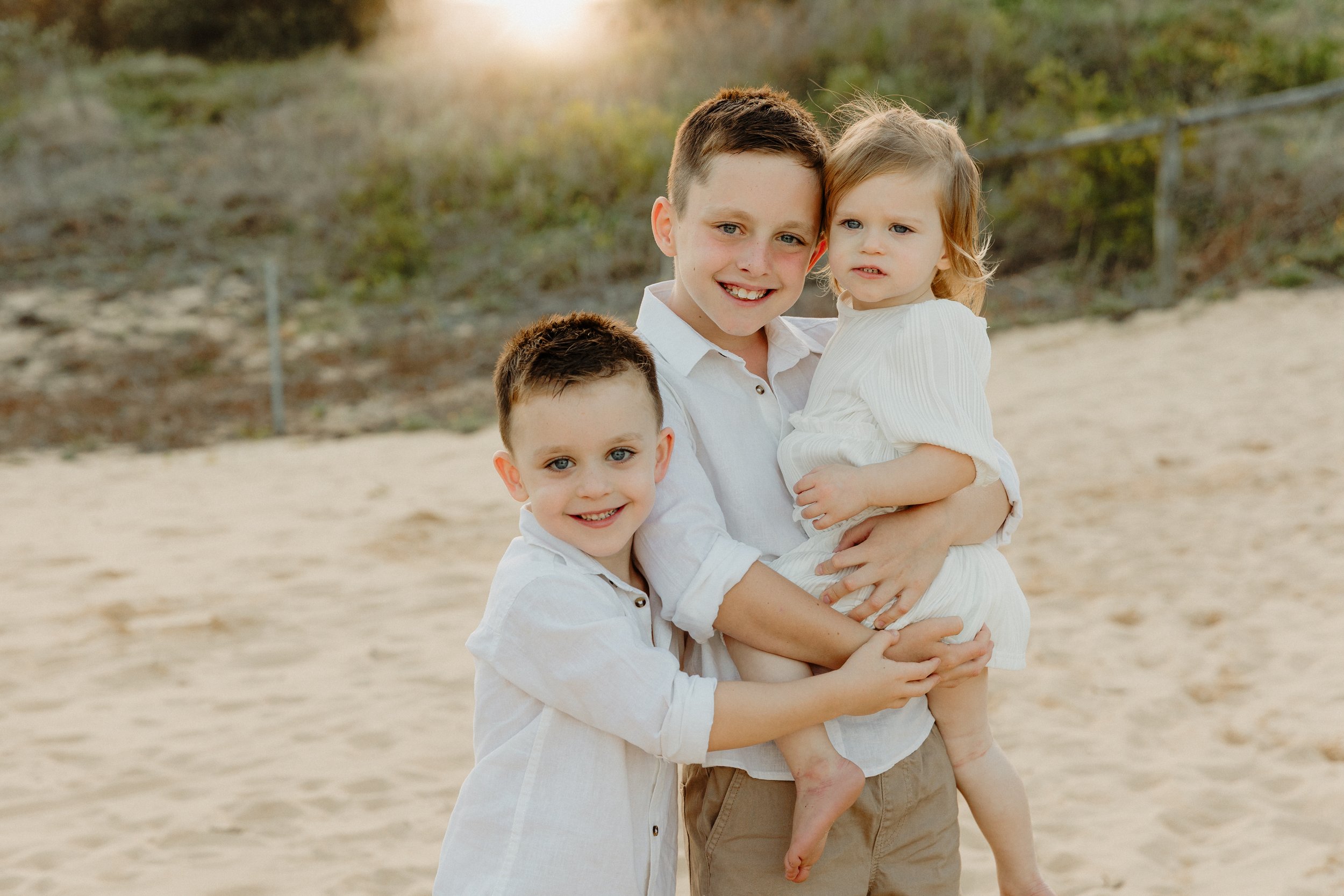 A smiling young man holding a young girl with blonde hair, and another young boy smiling, on a sandy beach with greenery in the background.