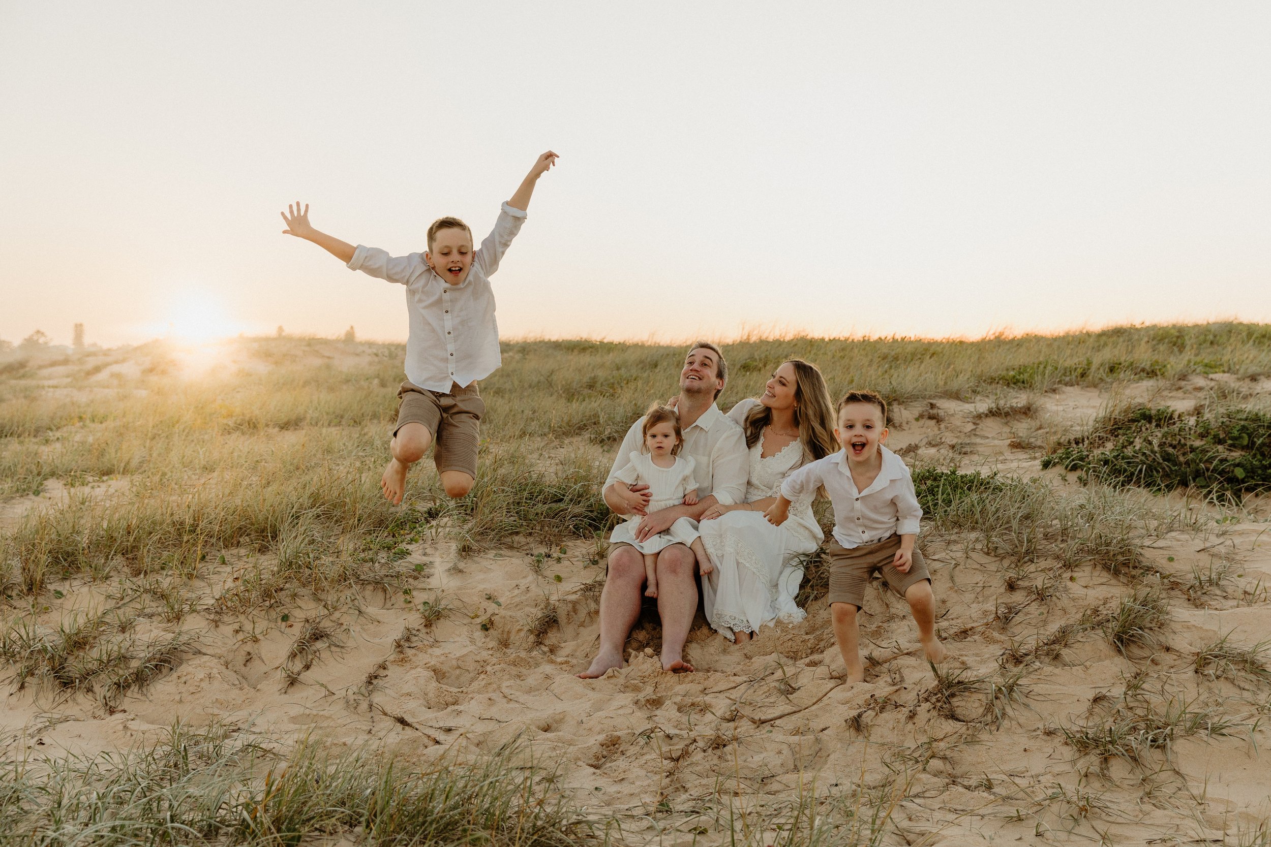 Happy family of five with two young children at the beach during sunset, with the youngest child jumping in the air.
