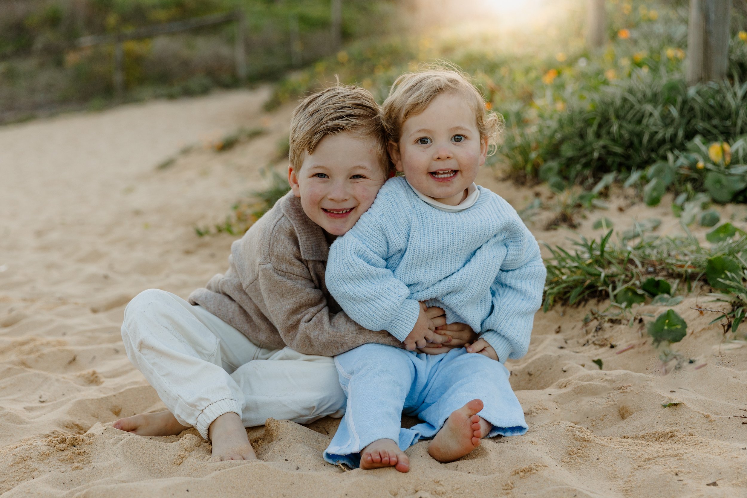 Two young children, a boy and a girl, sitting on sandy ground outdoors, smiling happily. The boy has short light brown hair and is wearing a beige sweater and white pants, hugging the girl from behind. The girl has curly blond hair, wearing a light b