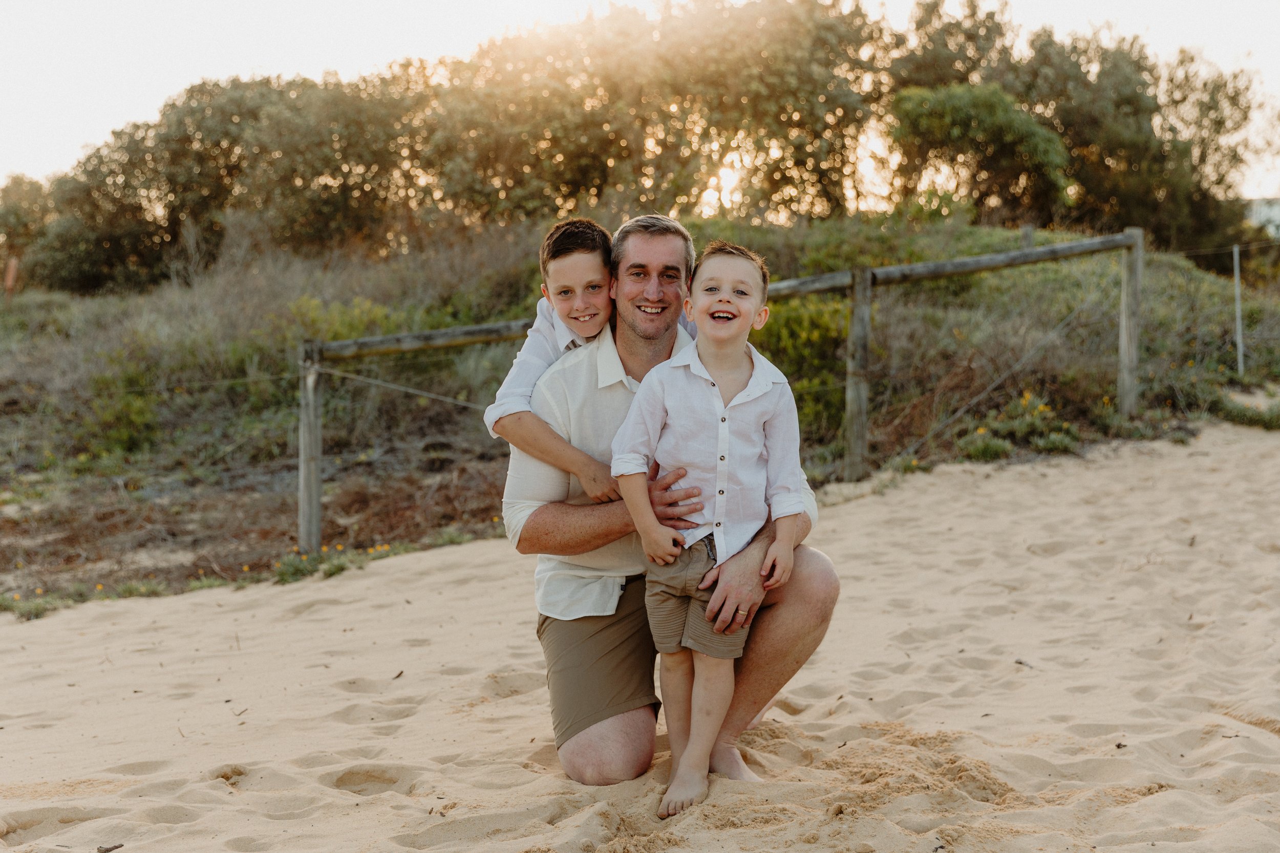 A man and two young boys on a sandy beach, smiling at the camera during sunset, with trees and a wooden fence in the background.