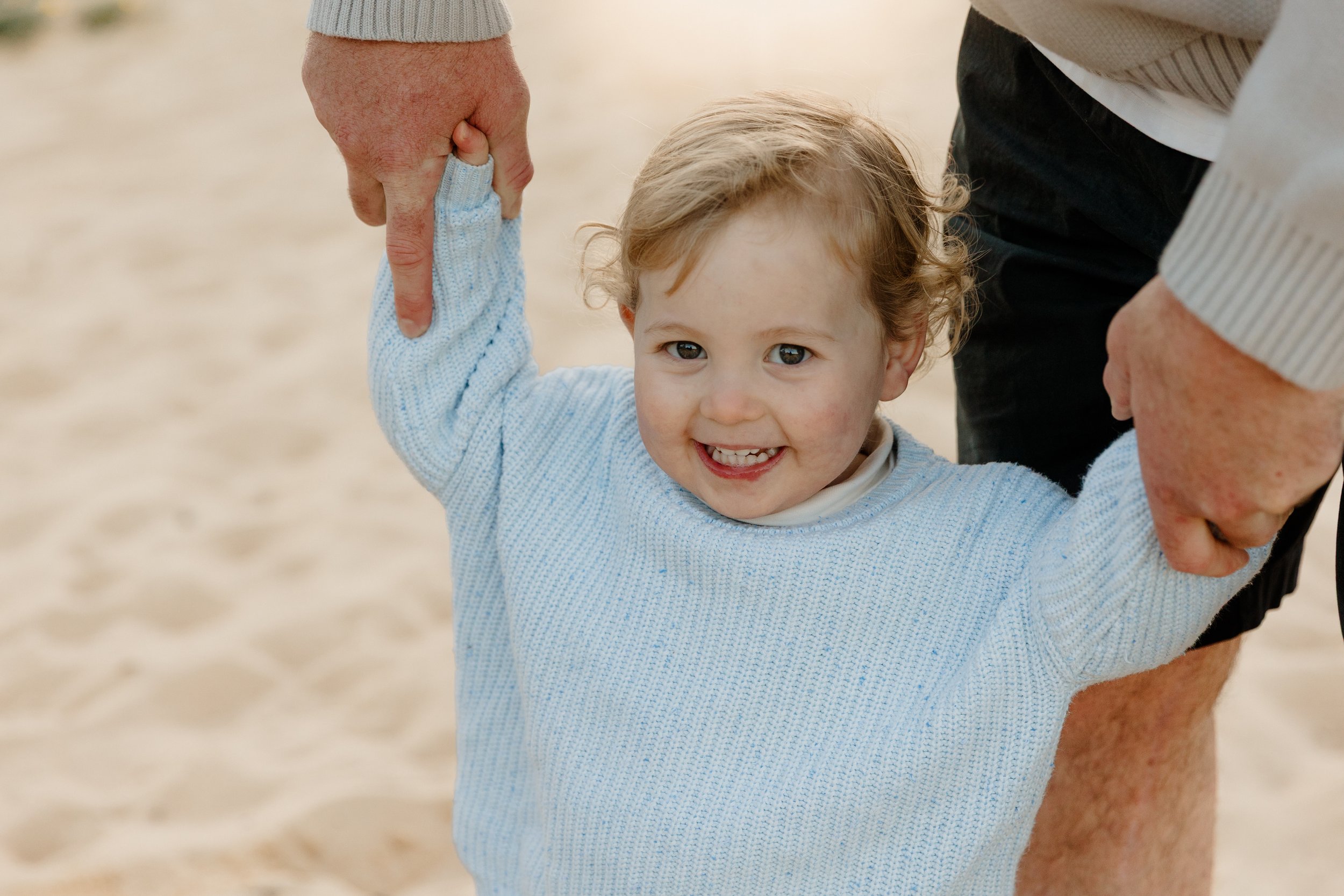 A young child smiling happily, being held by two adult hands, on a sandy beach.